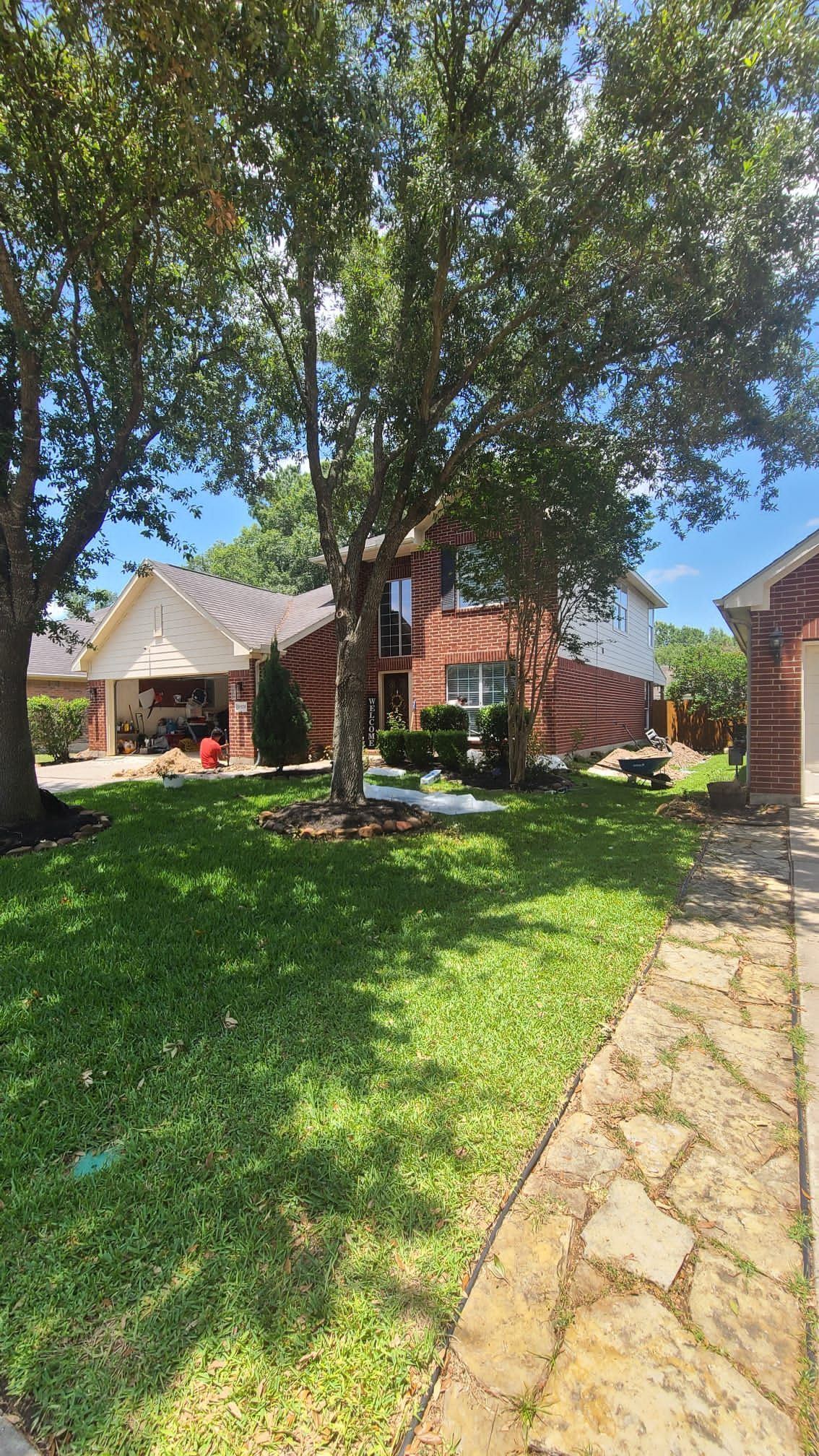Suburban front yard with large trees, green lawn, stone walkway, and brick houses in the background