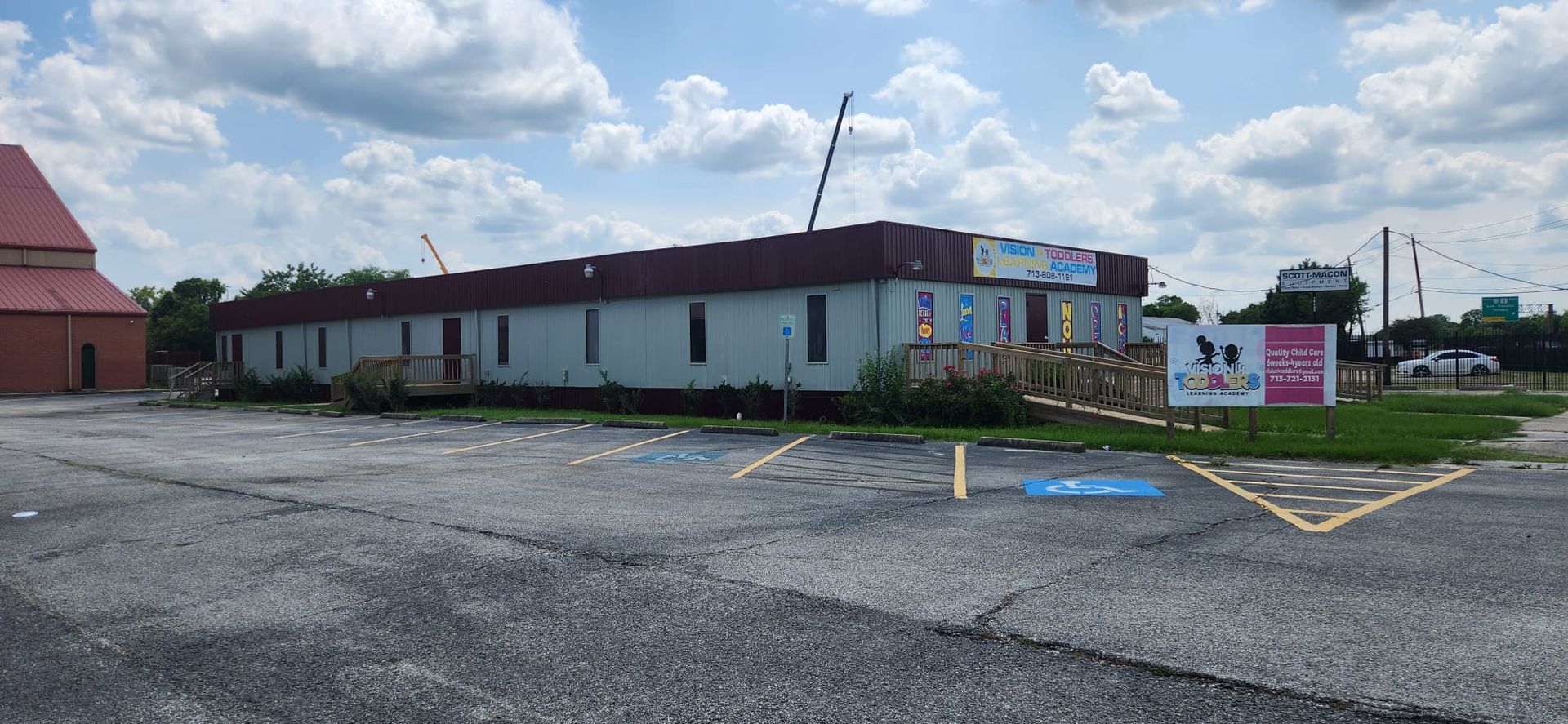 Low, long building with red roof beside an empty gravel lot under a partly cloudy sky.