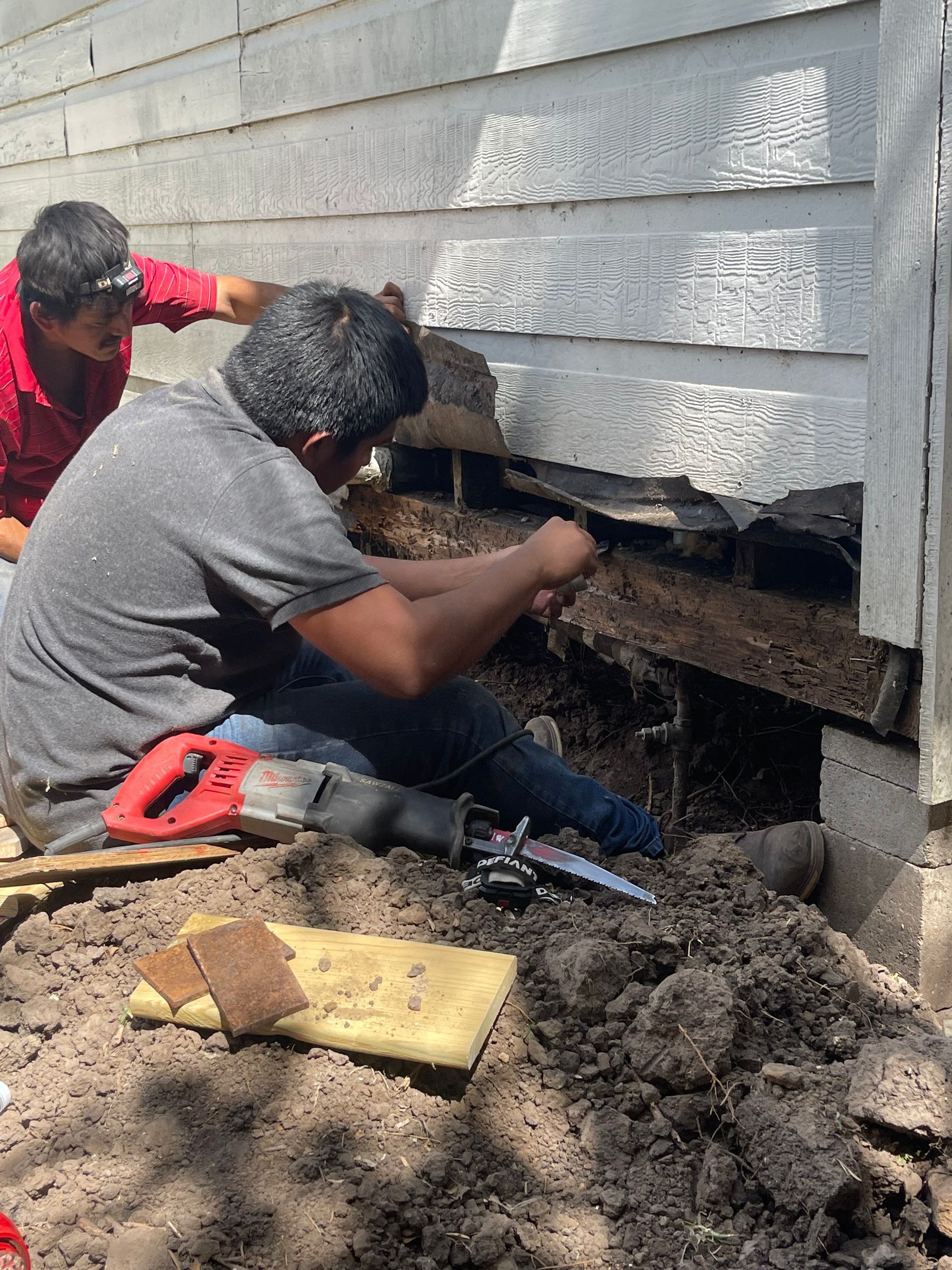 Workers digging under house siding to repair a foundation, with soil piled in the foreground.