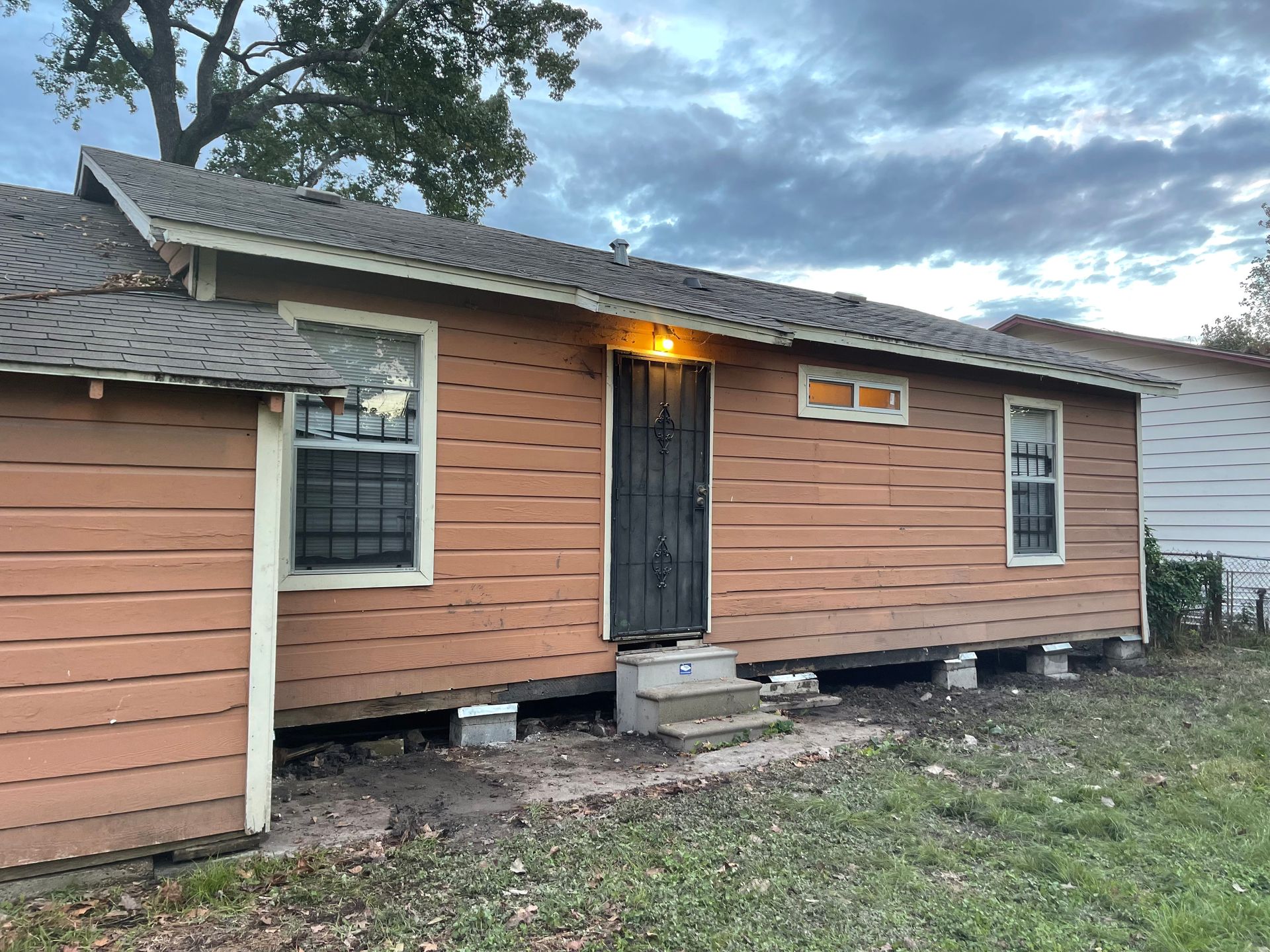 Small peach house with a black front door, lit porch light, and missing skirting on a cloudy day