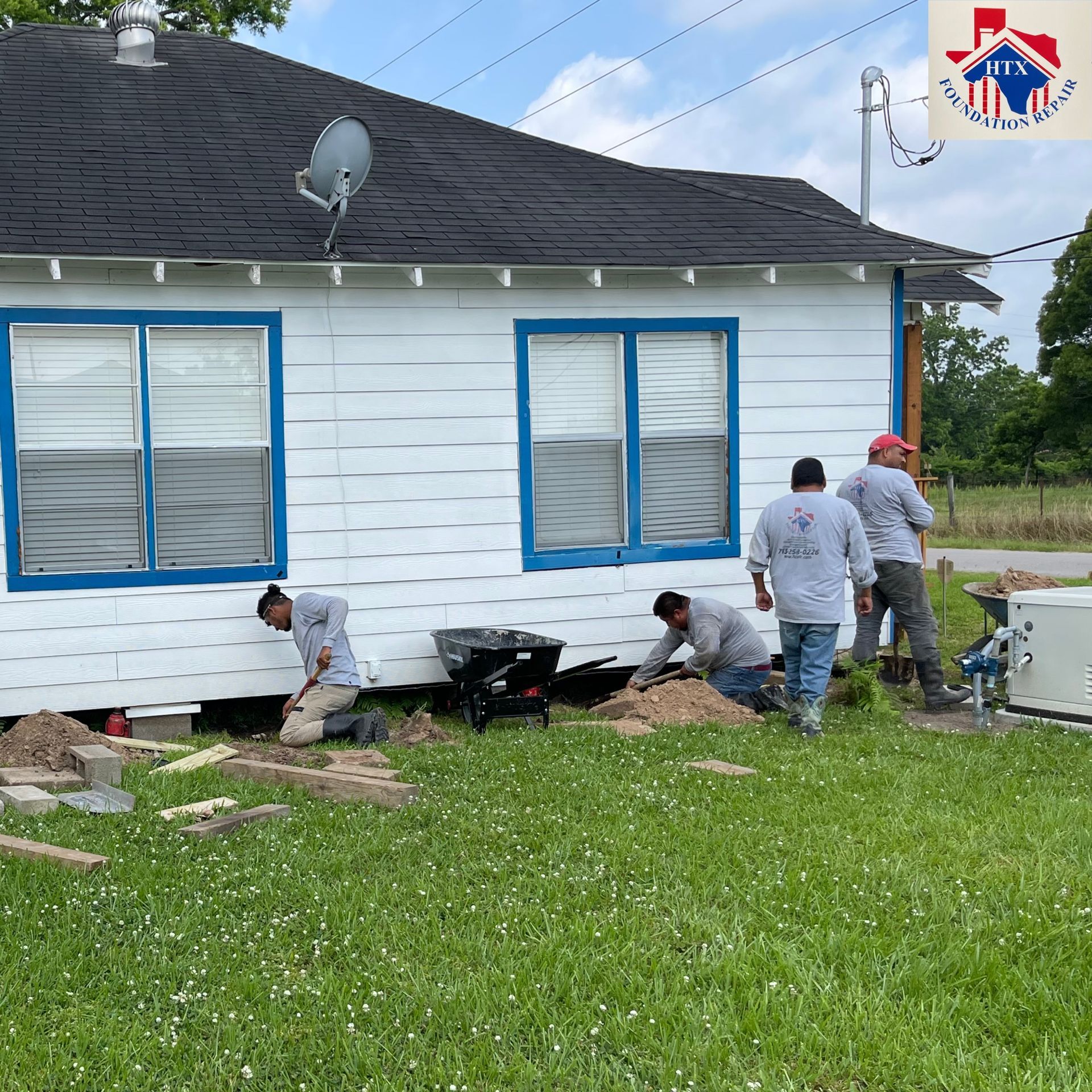 Workers repairing foundation outside a white house with blue-trimmed windows and a black roof