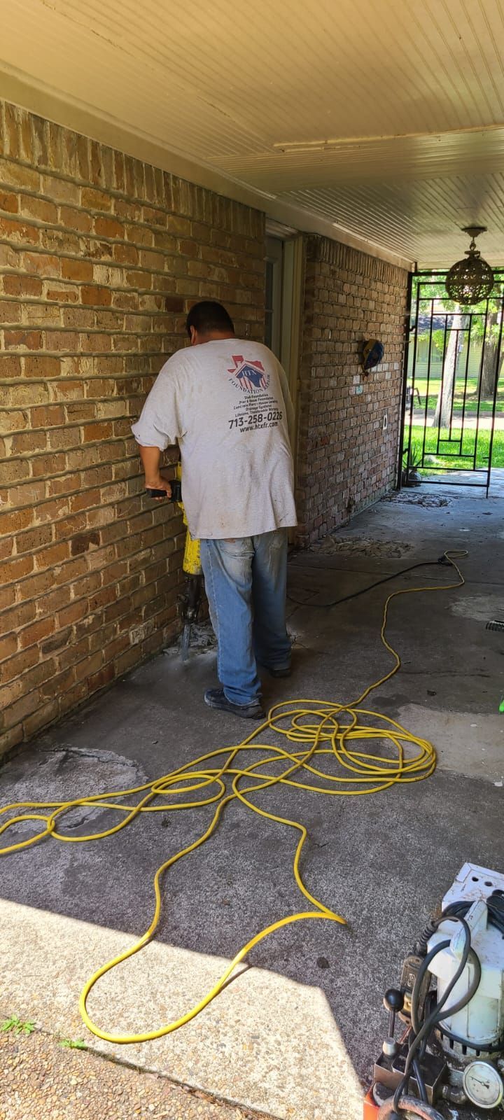Worker in white shirt installing or repairing something along a brick porch, with yellow cords on the floor.