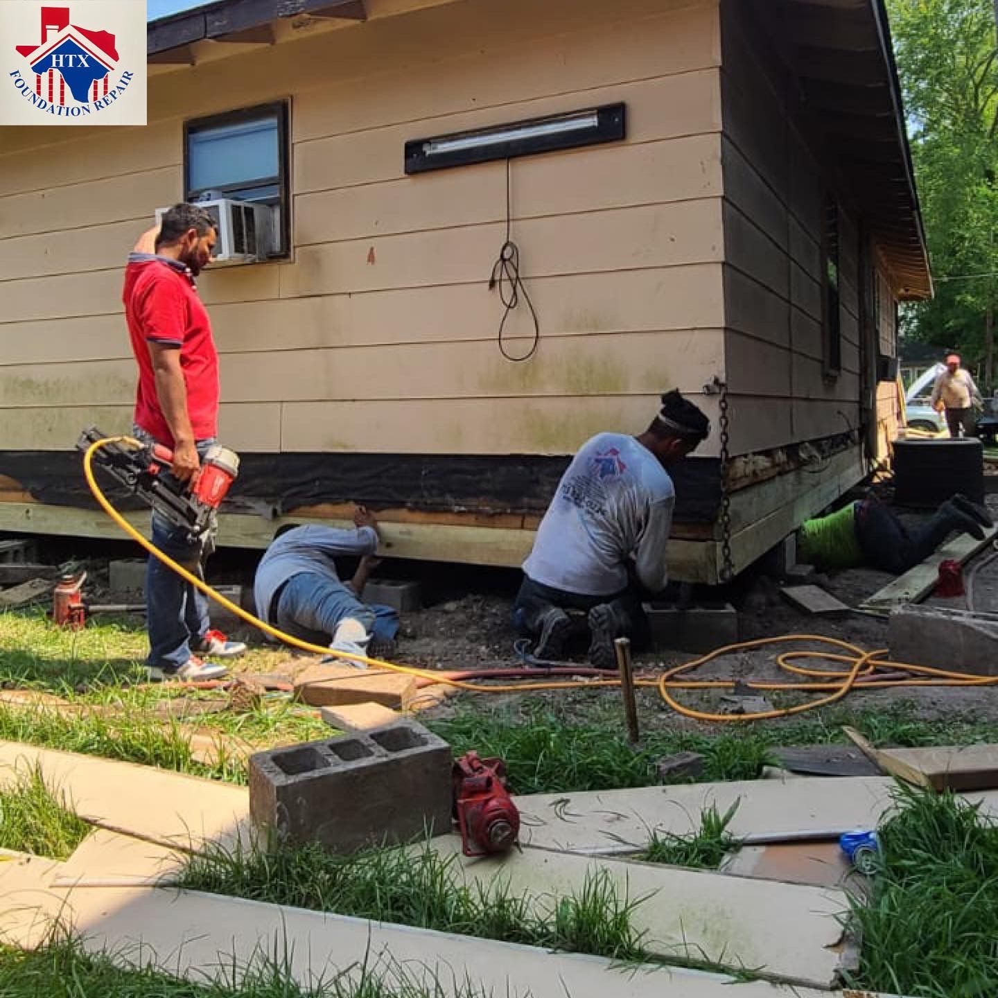 Workers lift a small tan house with jacks and hoses in a yard during foundation work