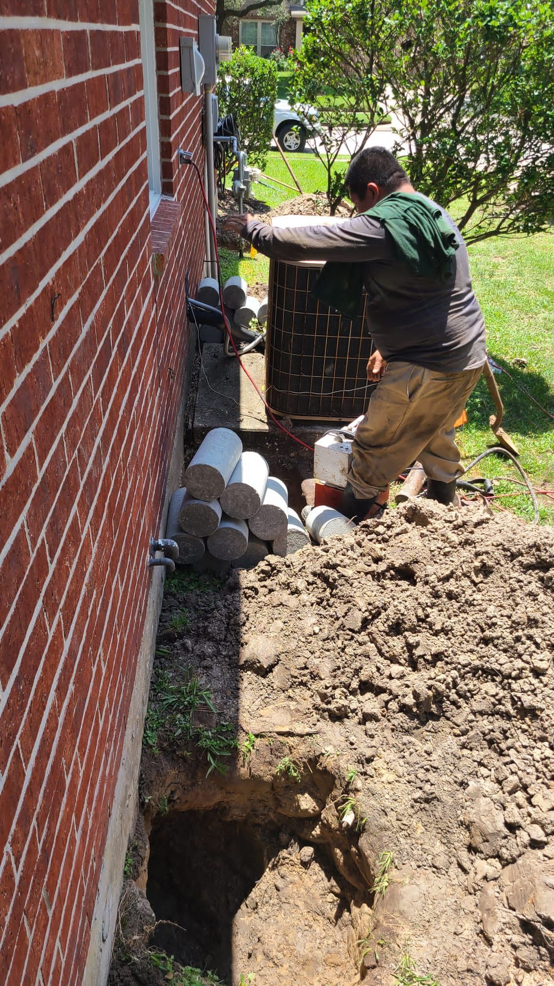 Worker digging beside a brick house, moving rocks near a foundation trench in a yard.