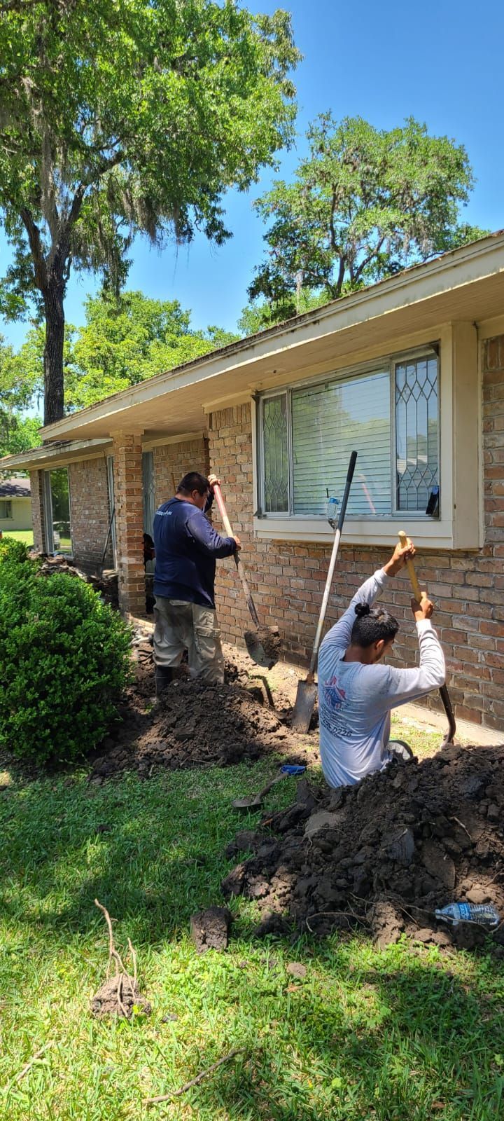 Two workers shoveling dirt beside a house with a green lawn and trees in the yard.