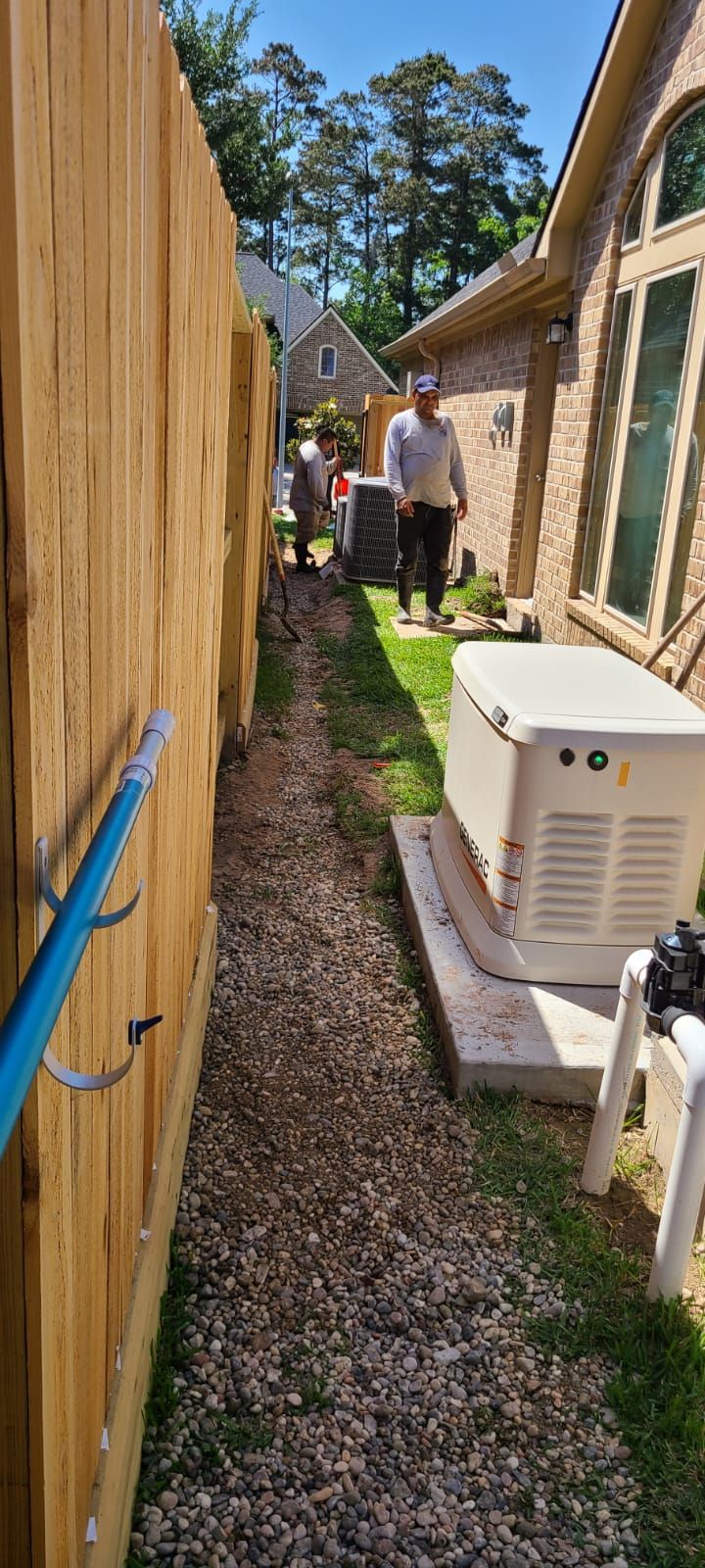 Narrow side yard with gravel path, wooden fence, house wall, HVAC unit, and a person in the distance.
