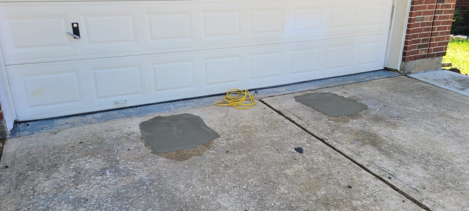 Concrete driveway in front of a white garage door with two dark stains and a yellow hose near the wall