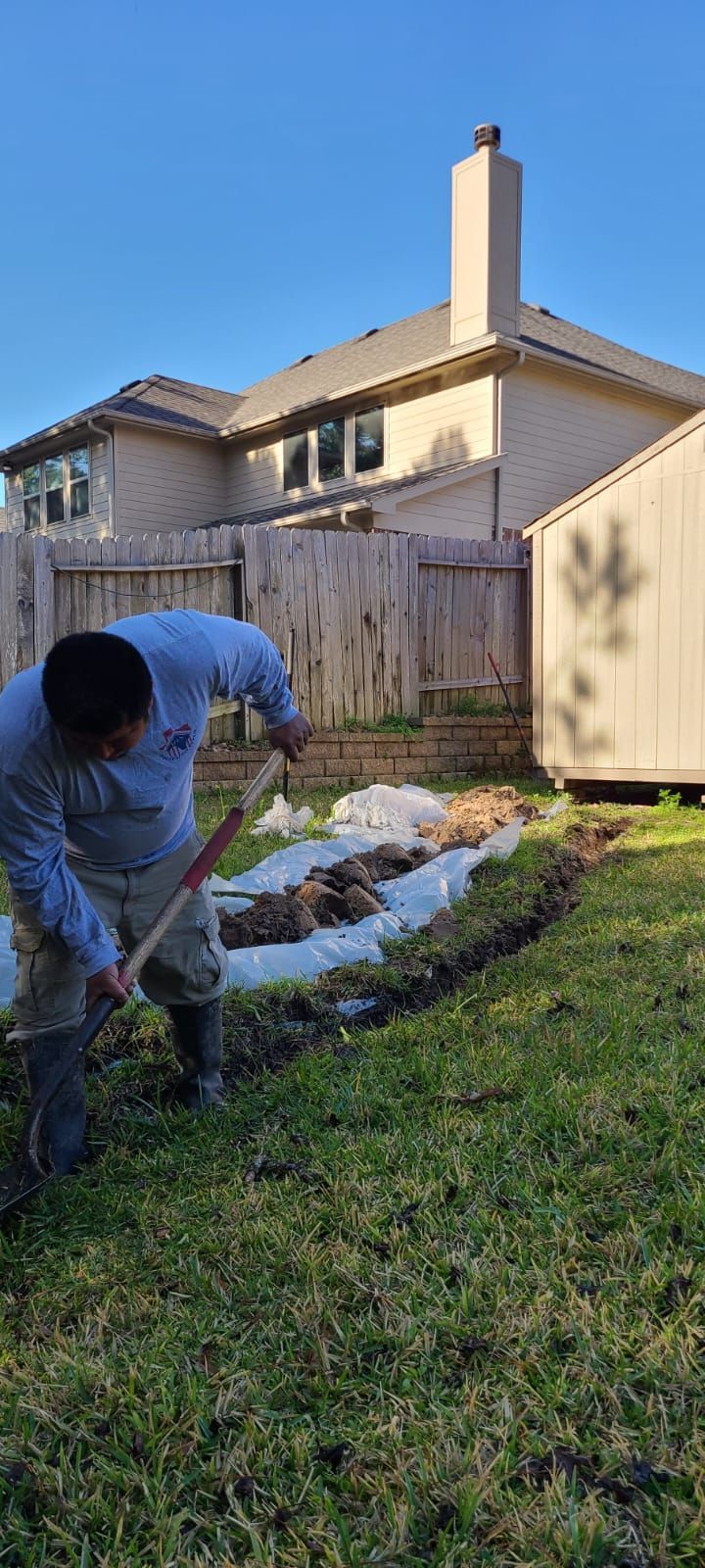 Person digging in a backyard garden beside a house, with a fence and bright blue sky.