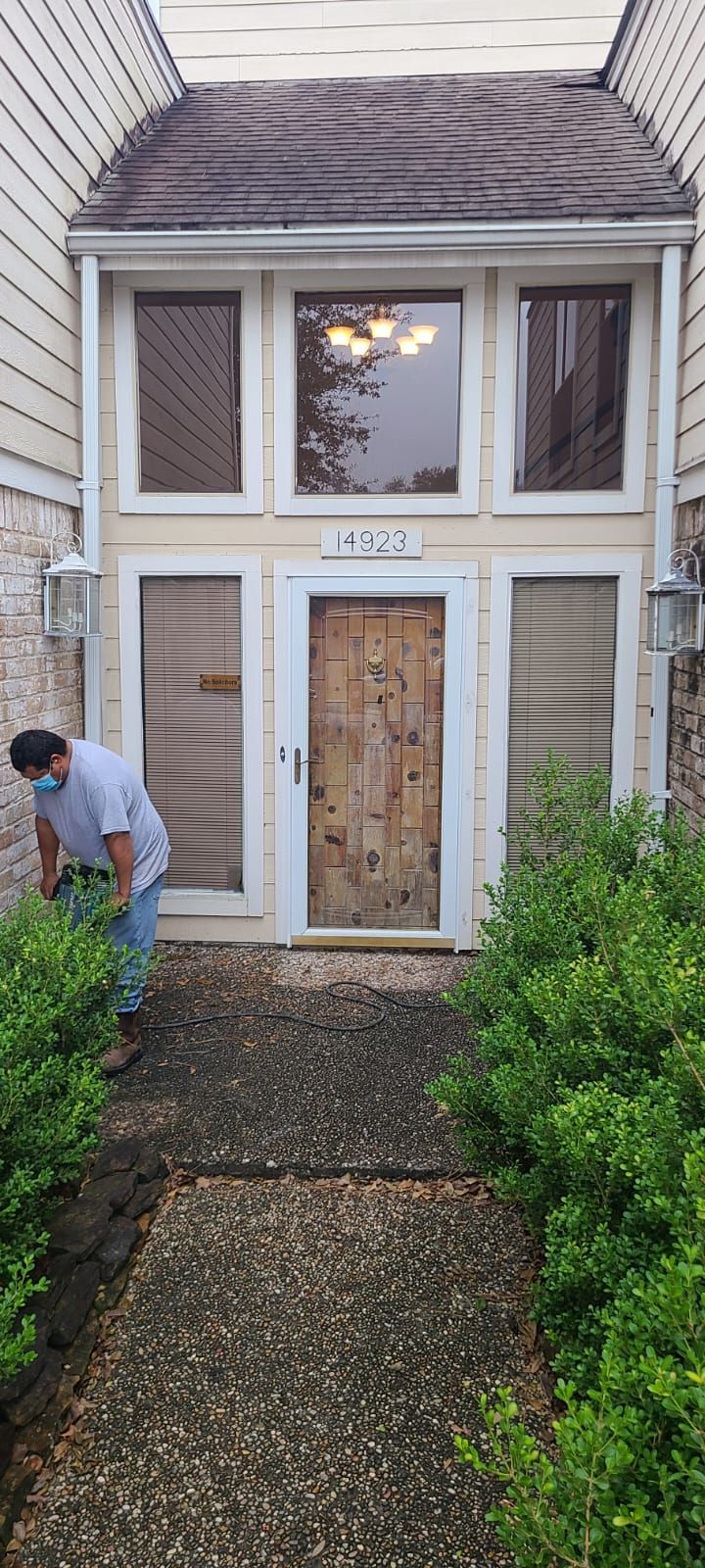 Front view of a house with boarded windows and door, stone path, and a person trimming bushes on the left.