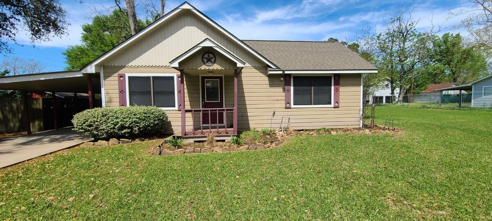 Single-story beige house with a front porch, pink-trimmed windows, and a large green lawn.