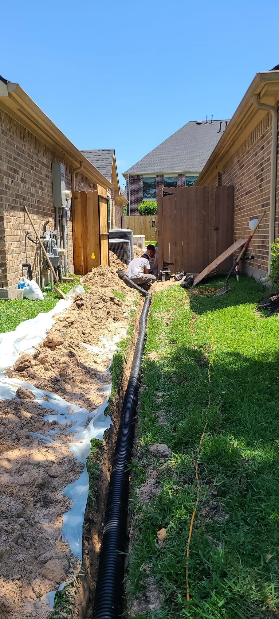 Narrow muddy alley between wooden houses with snow patches and green grass under a clear blue sky