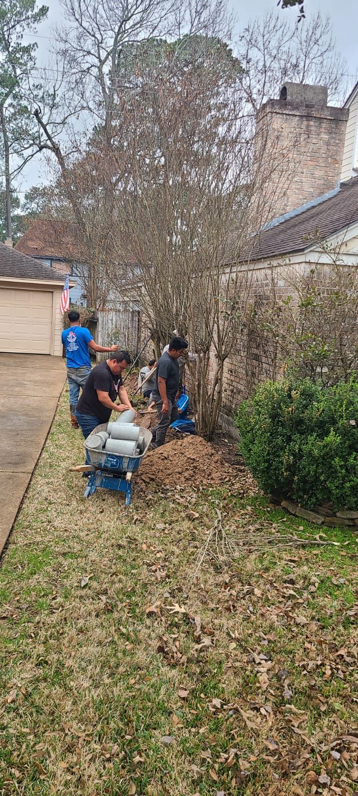 People digging in a leaf-covered backyard beside a house and bushes under cloudy skies