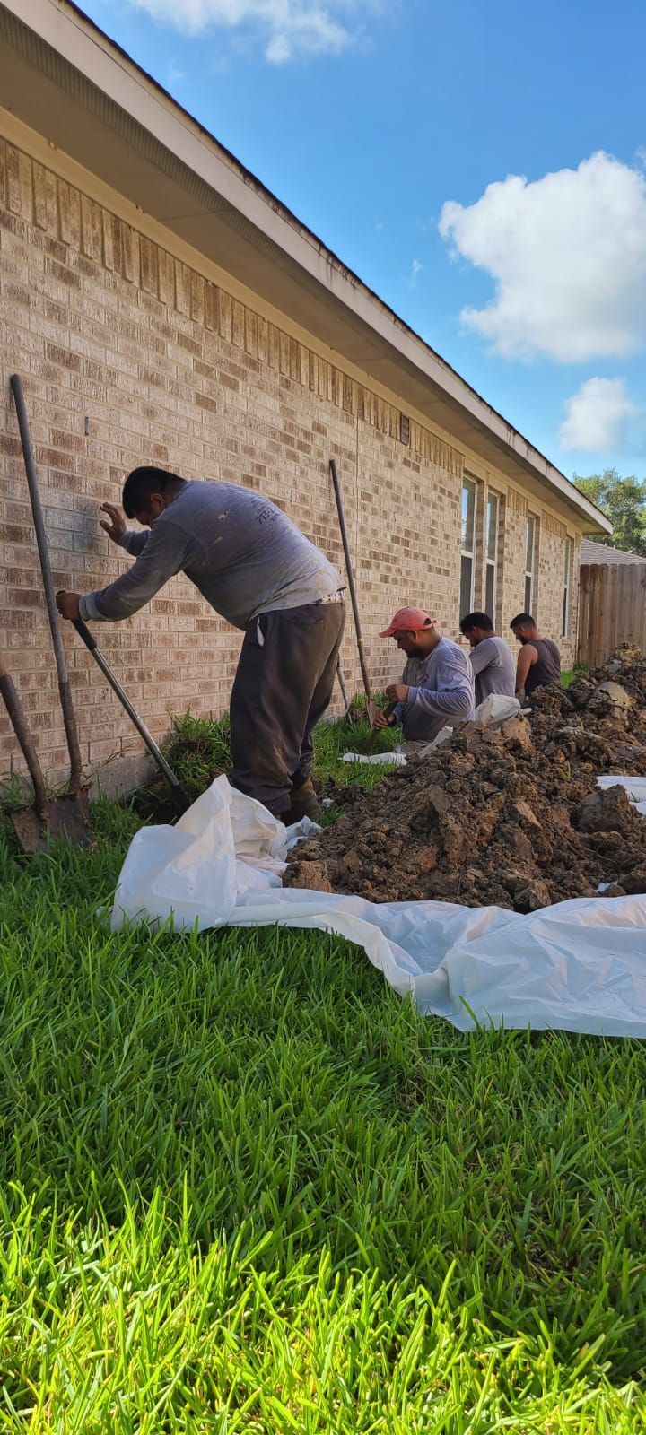 People stacking soil against a long brick wall beside a grassy field under a blue sky.
