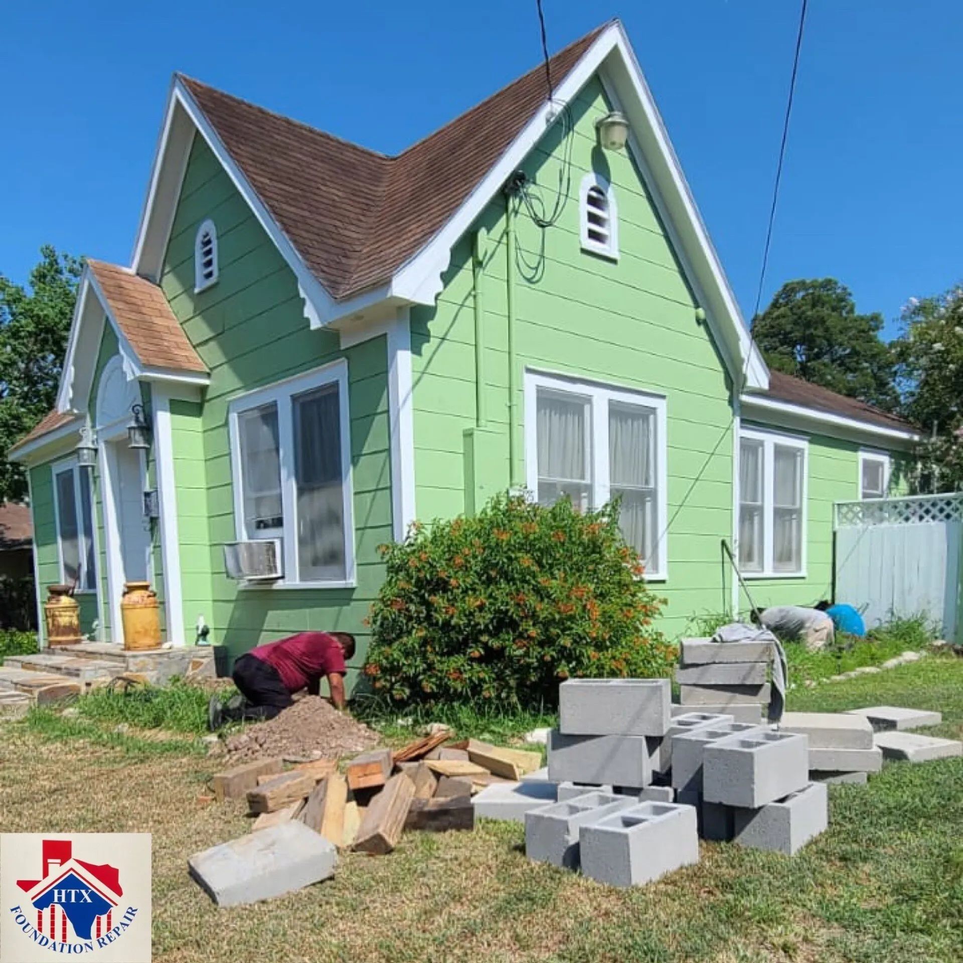 Green house with workers laying cinder blocks in the yard and a pile of bricks nearby