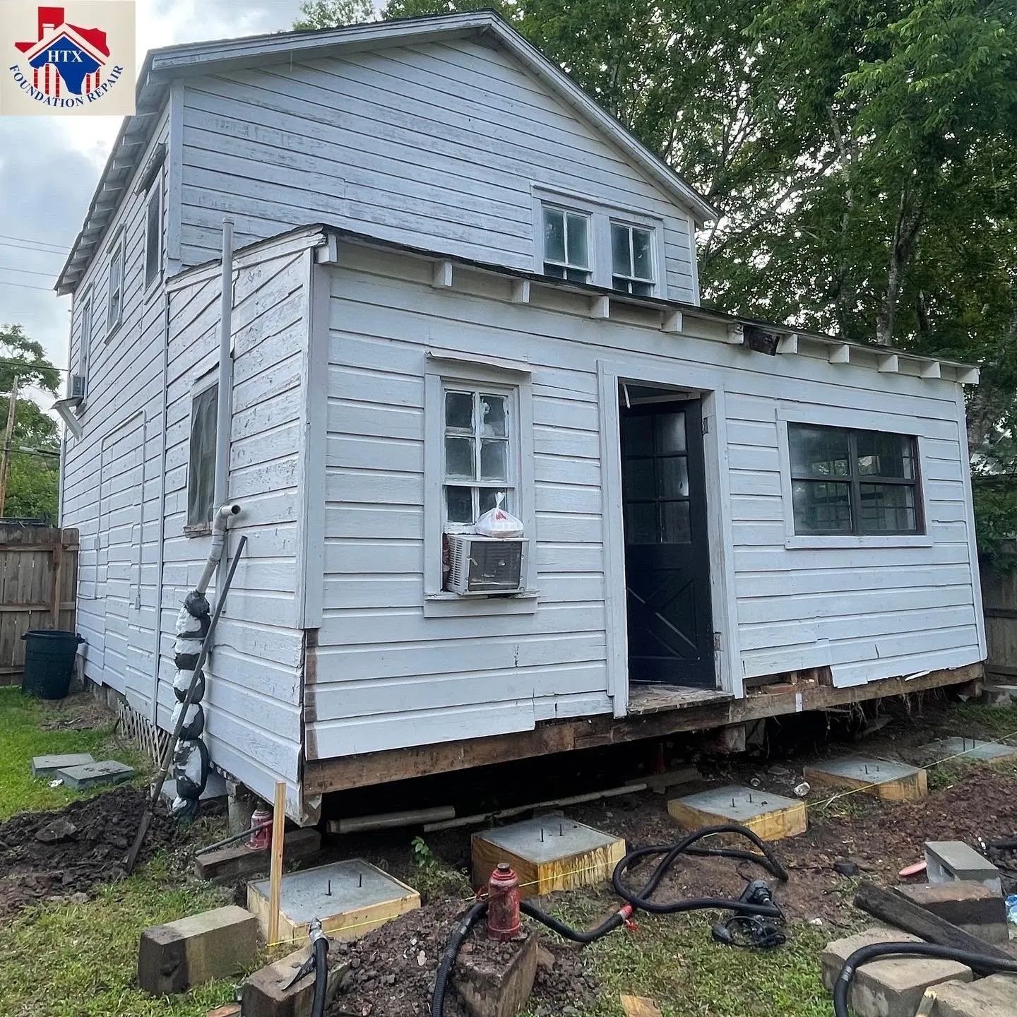 Partially raised white house on blocks with open door and windows in a yard under repair