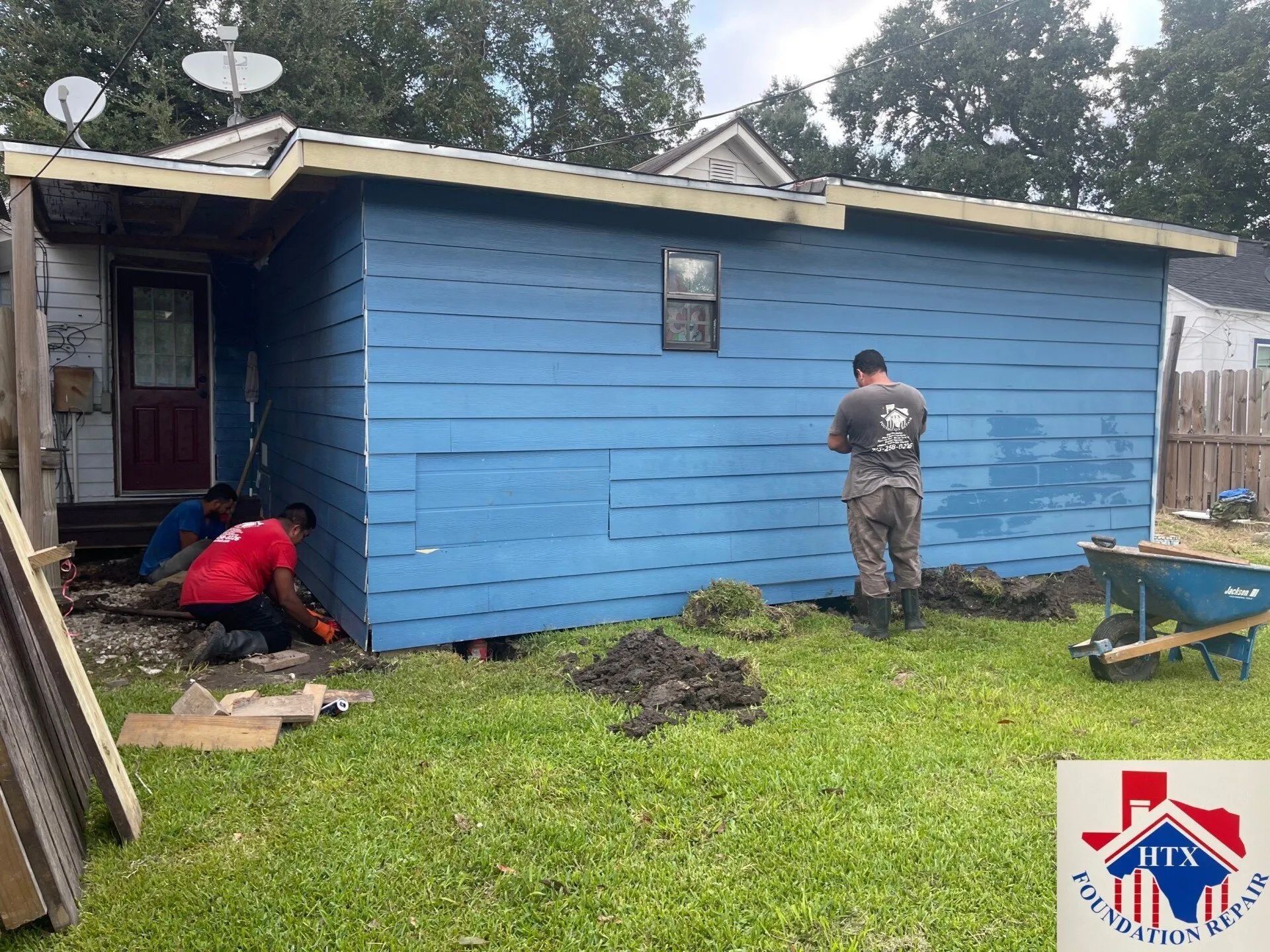 Two people working outside a blue house, one kneeling by the door and one standing in the yard.
