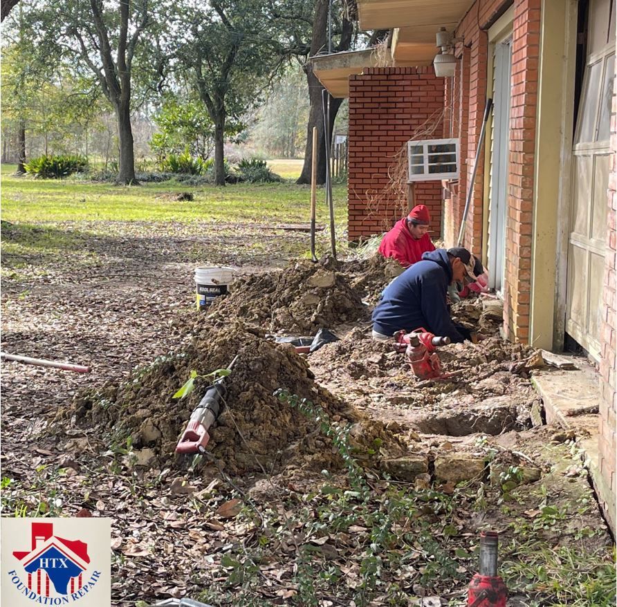 Two workers kneel by a brick house, digging and clearing mud along the foundation after heavy rain.