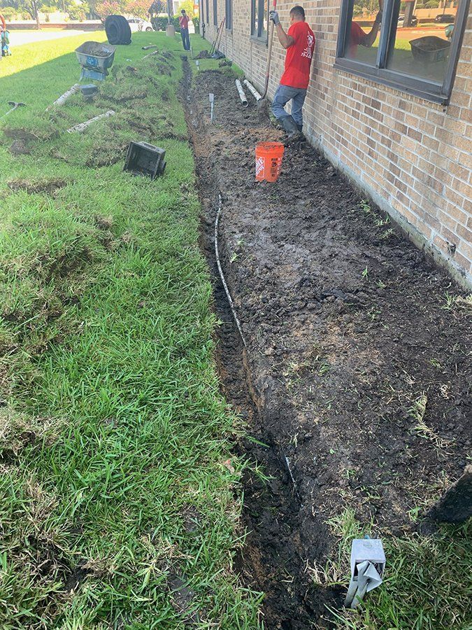 Worker digging a trench beside a brick building, with a bucket and tools on the ground.