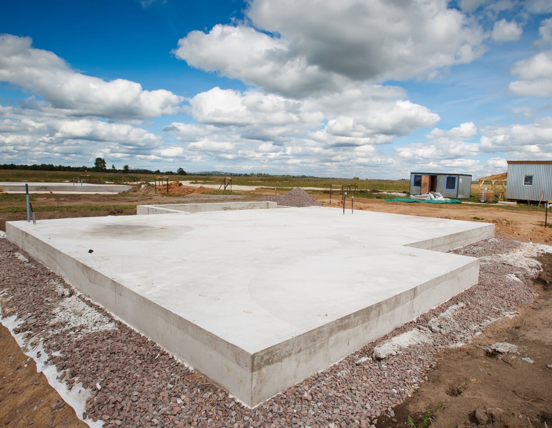 Concrete foundation slab at a construction site under a cloudy sky, with gravel and small site buildings nearby