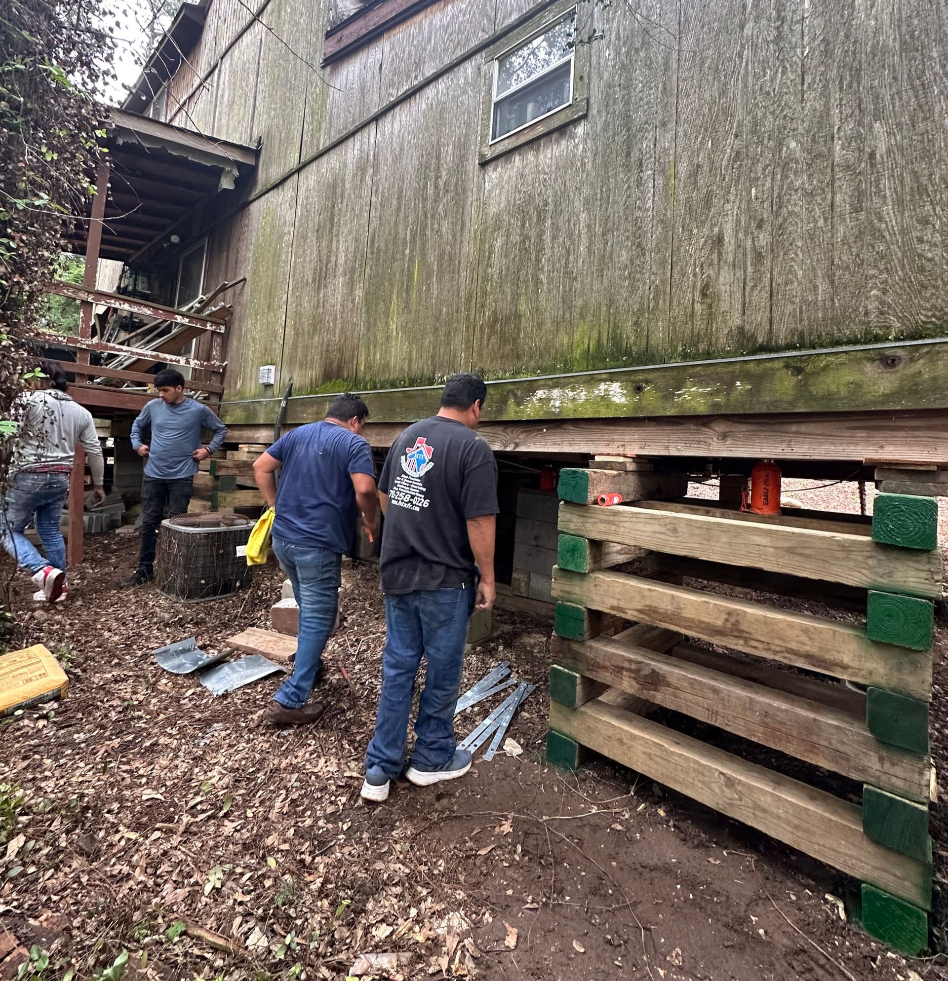People inspecting a weathered wooden building with stacked pallets beside it in a muddy outdoor area