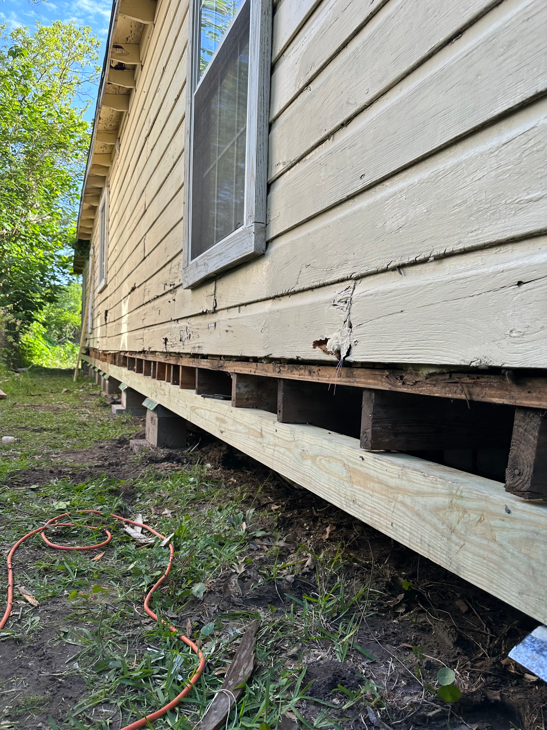 Weathered house siding lifted above exposed foundation supports beside a dirt yard and trees.