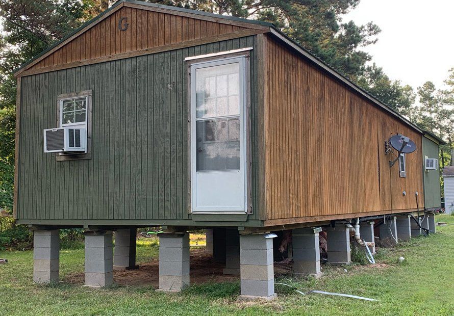 Elevated wooden house on concrete piers with green and brown siding, small window air conditioner, and a white door.