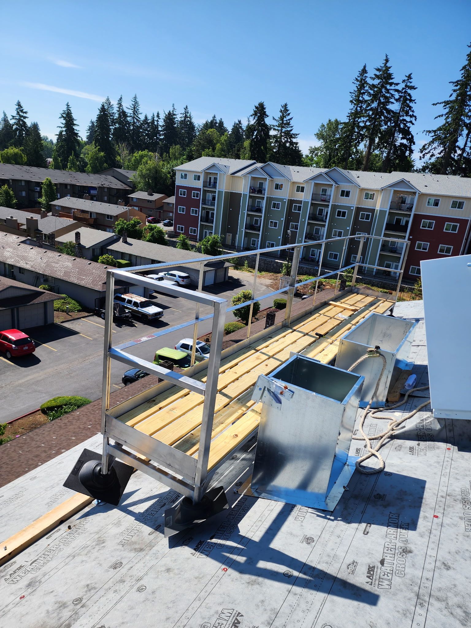 A metal rolling scaffolding platform sits on a flat roof overlooking an apartment complex on a sunny day.