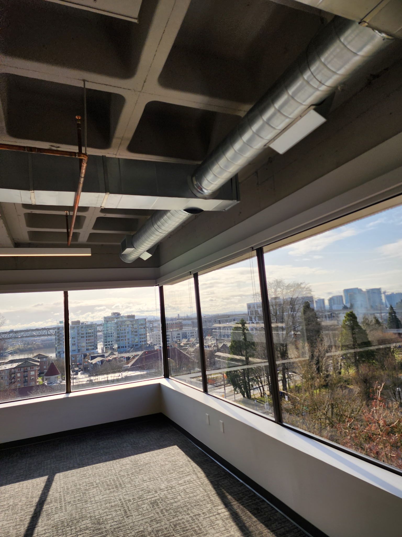 Office interior with large windows, city view, gray ceiling with ducts, and light-colored carpet.
