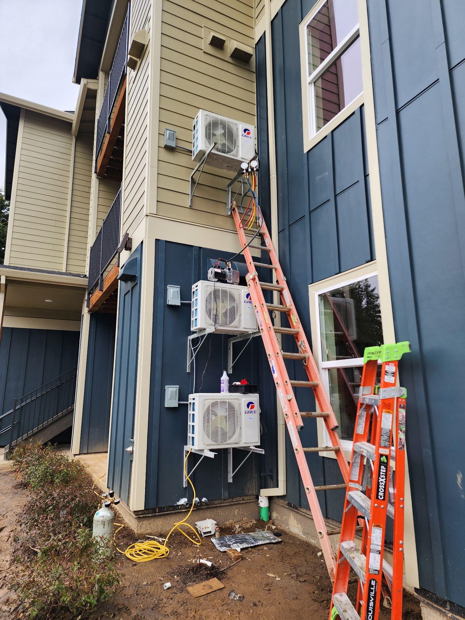 HVAC units mounted on a blue-sided building with a ladder beside them; wires and tools are on the ground.
