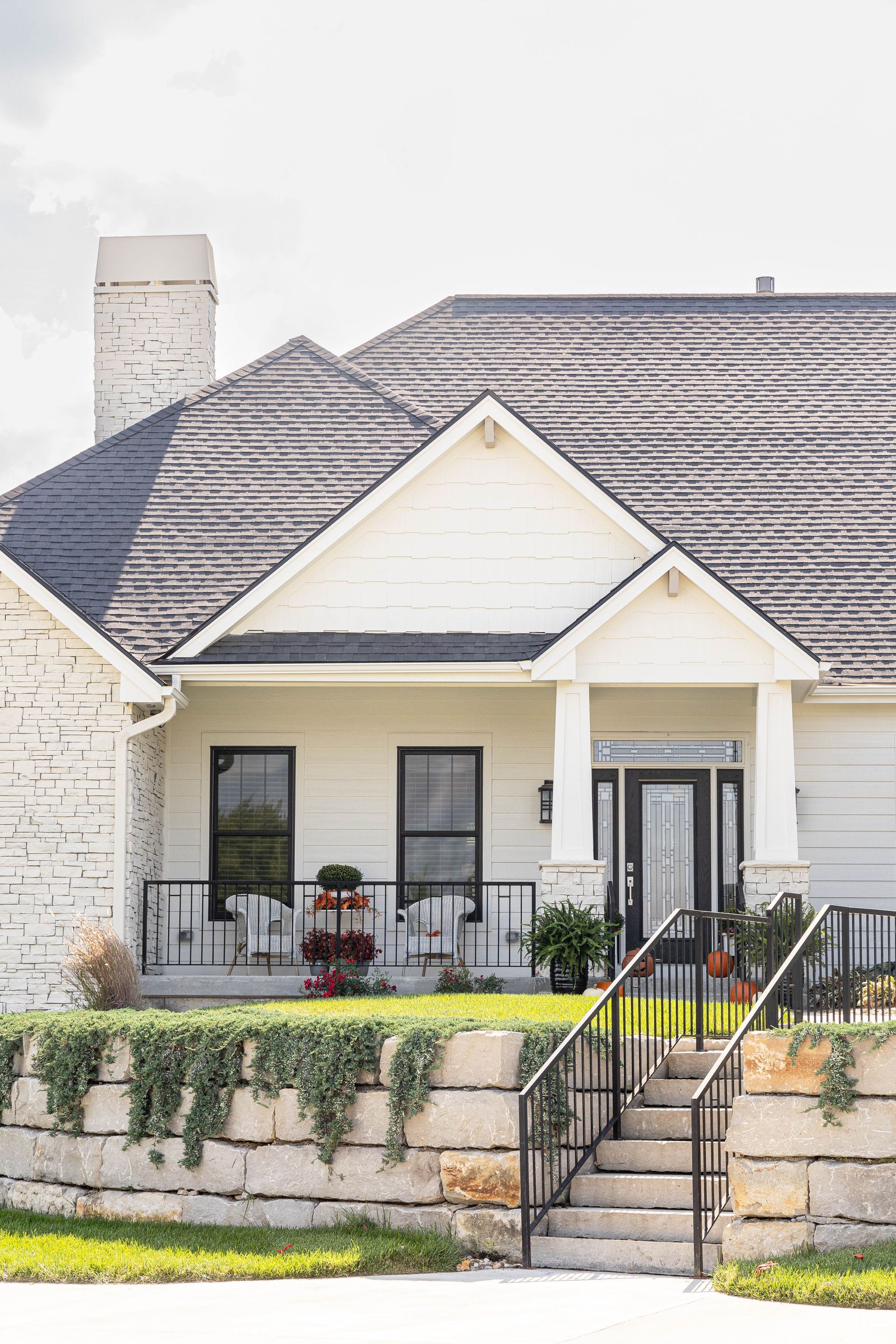 A white house with a gray roof and a stone wall in front of it.