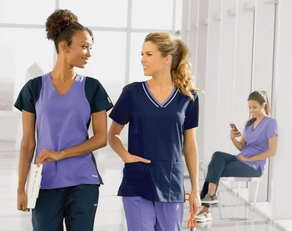 Two people in scrubs walking and talking in a medical setting, one person seated and using a phone.