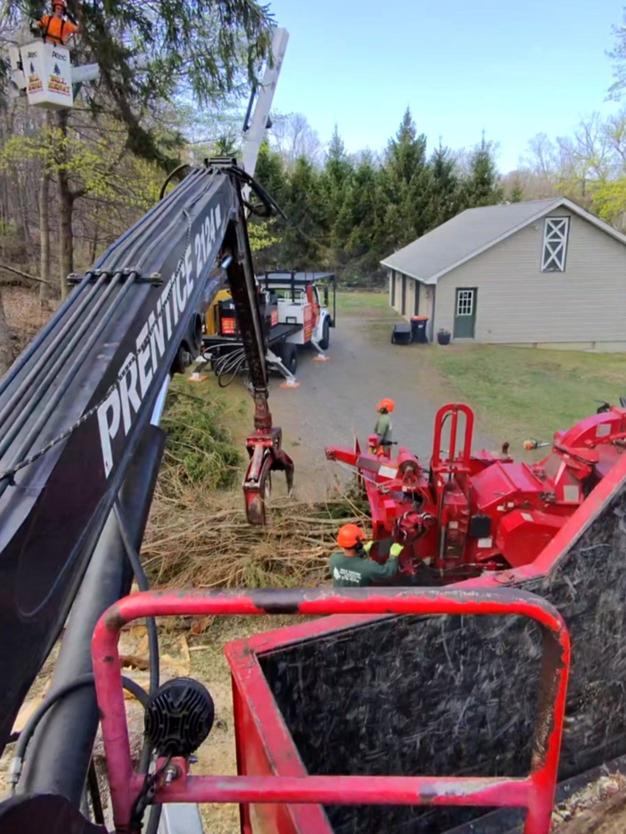 Crane lifting a red tractor attachment in a grassy yard beside a beige shed and trees.