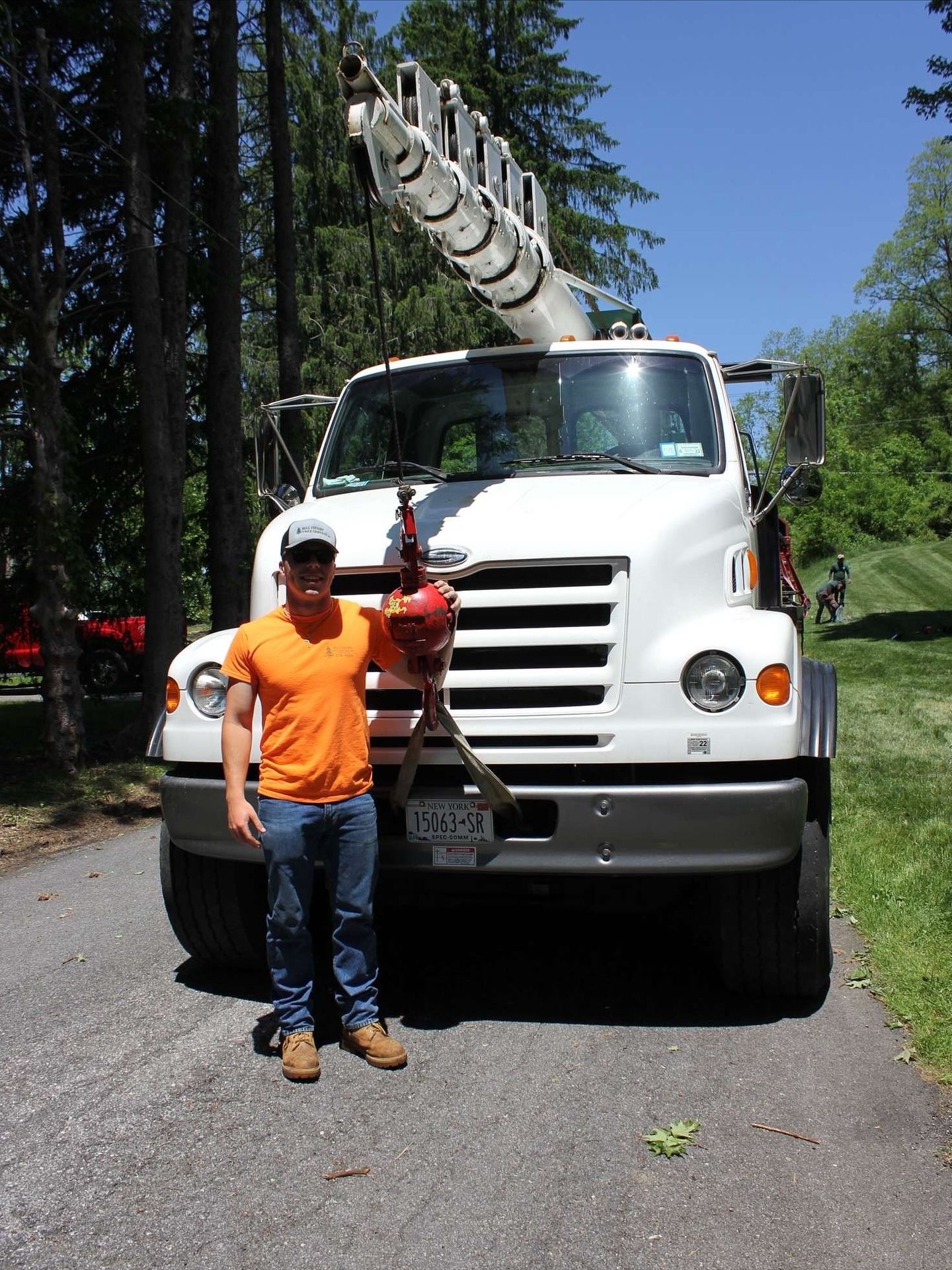 Man standing in front of a white utility truck with a ladder on top on a tree-lined road.