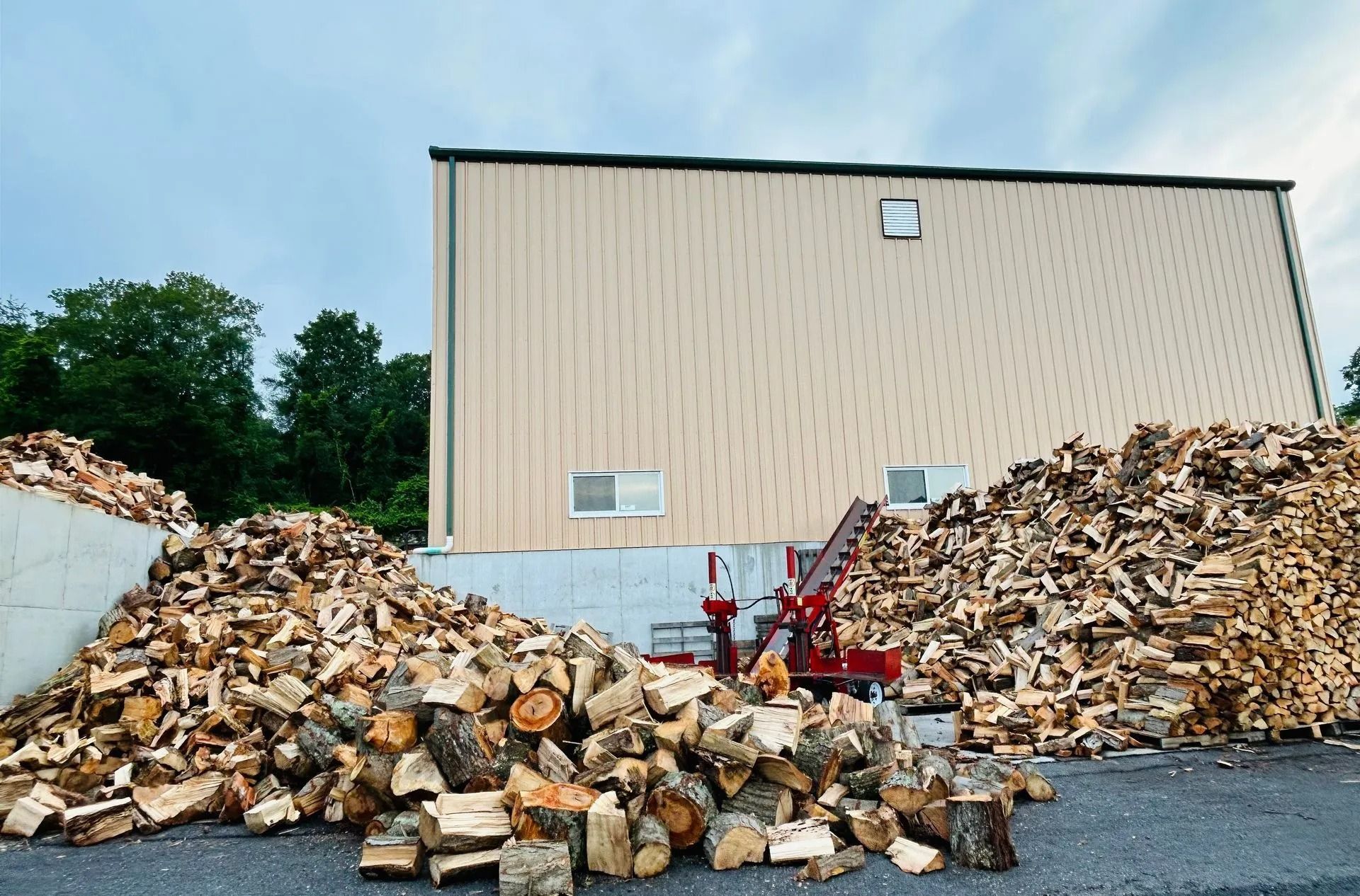 Large wood piles beside a beige industrial building on a gravel lot