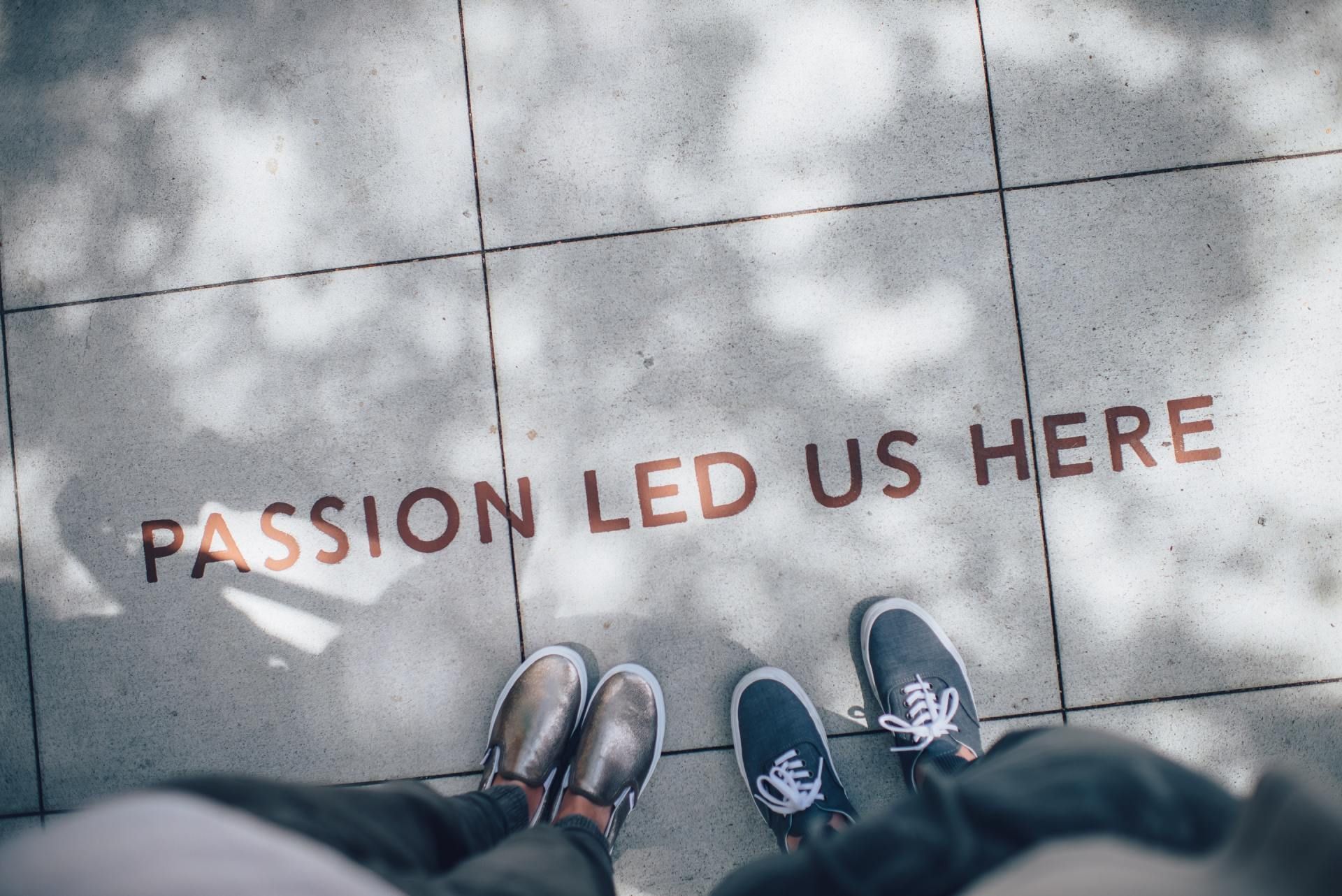 Feet standing on pavement beside the words “PASSION LED US HERE” painted in red.