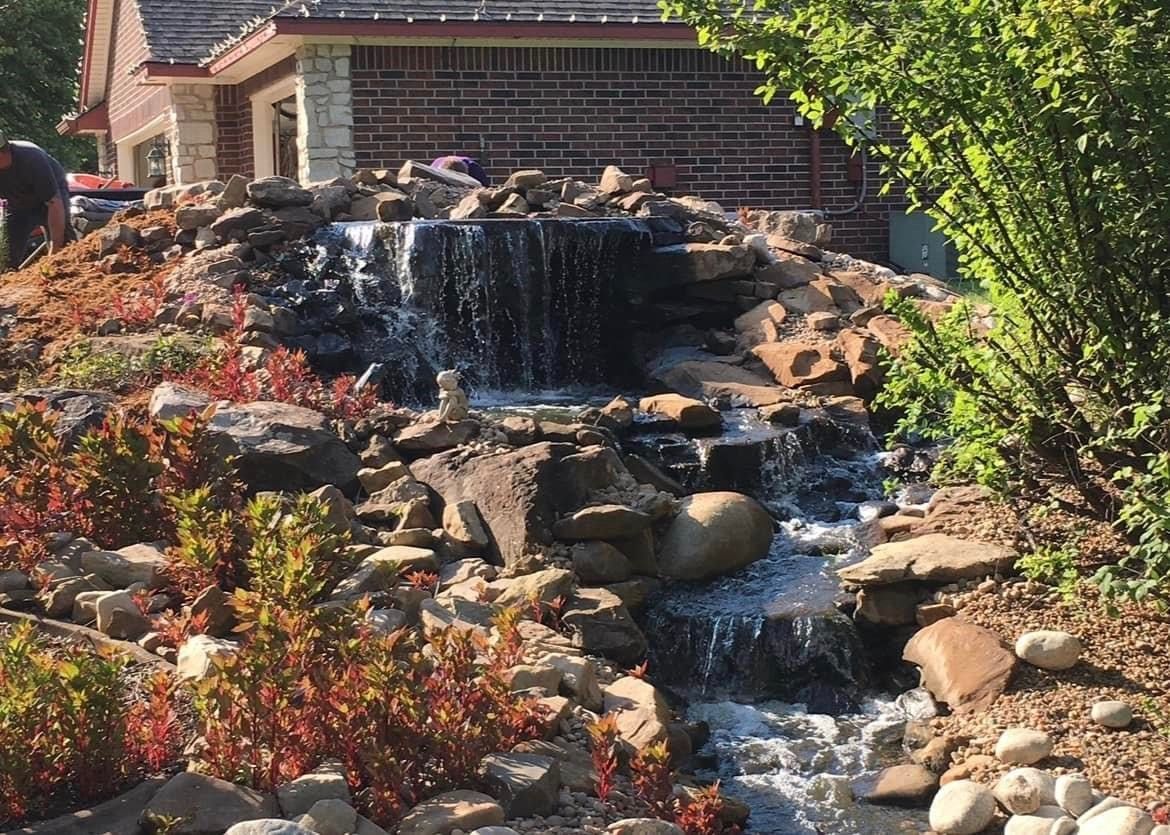 A waterfall is surrounded by rocks and plants in front of a house.