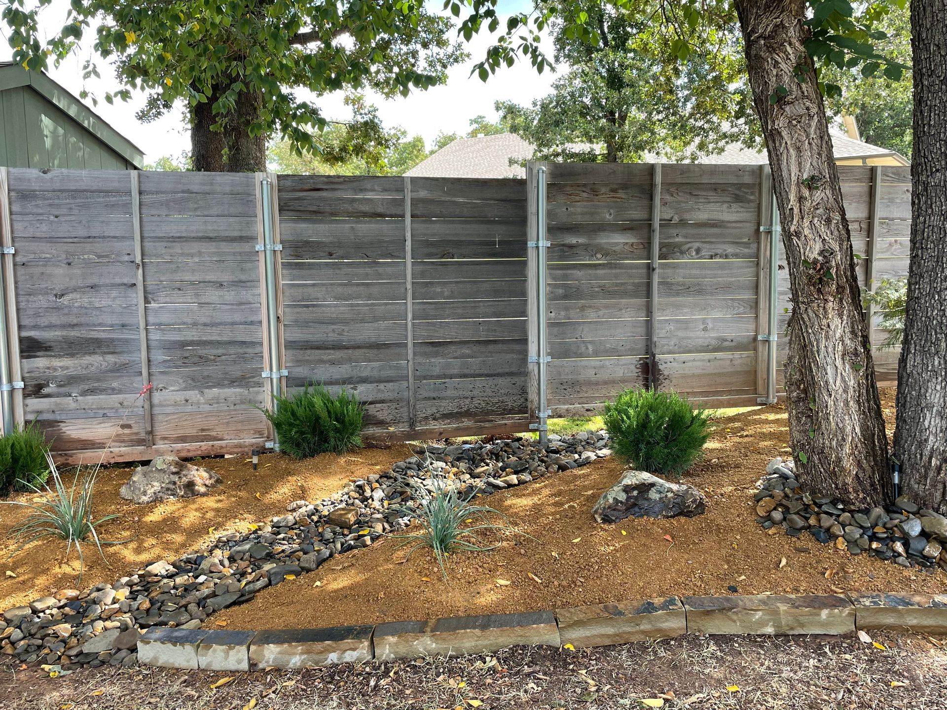 A wooden fence is surrounded by trees and rocks in a yard.