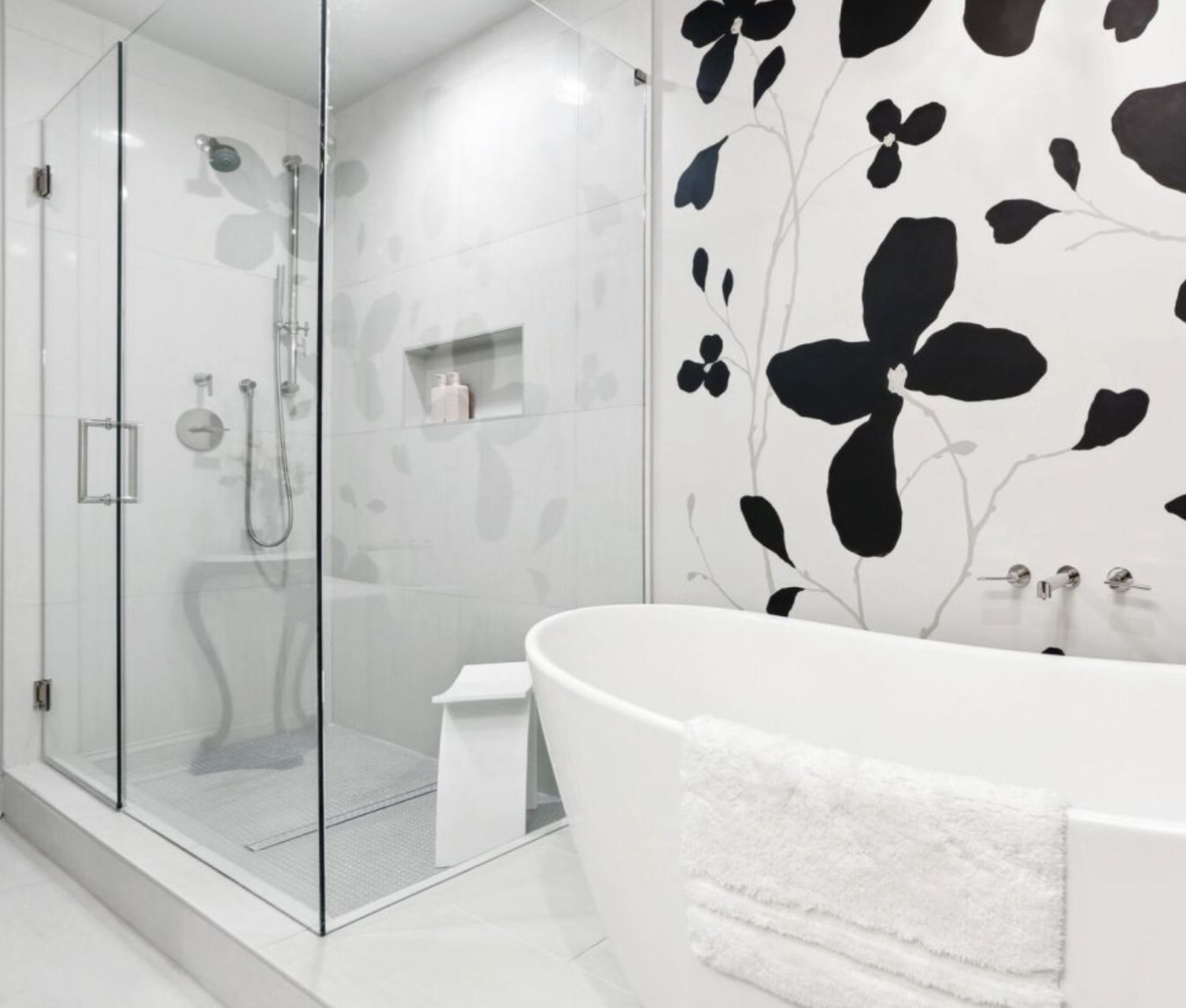Modern white bathroom with glass shower, large black floral wall, and a white bathtub