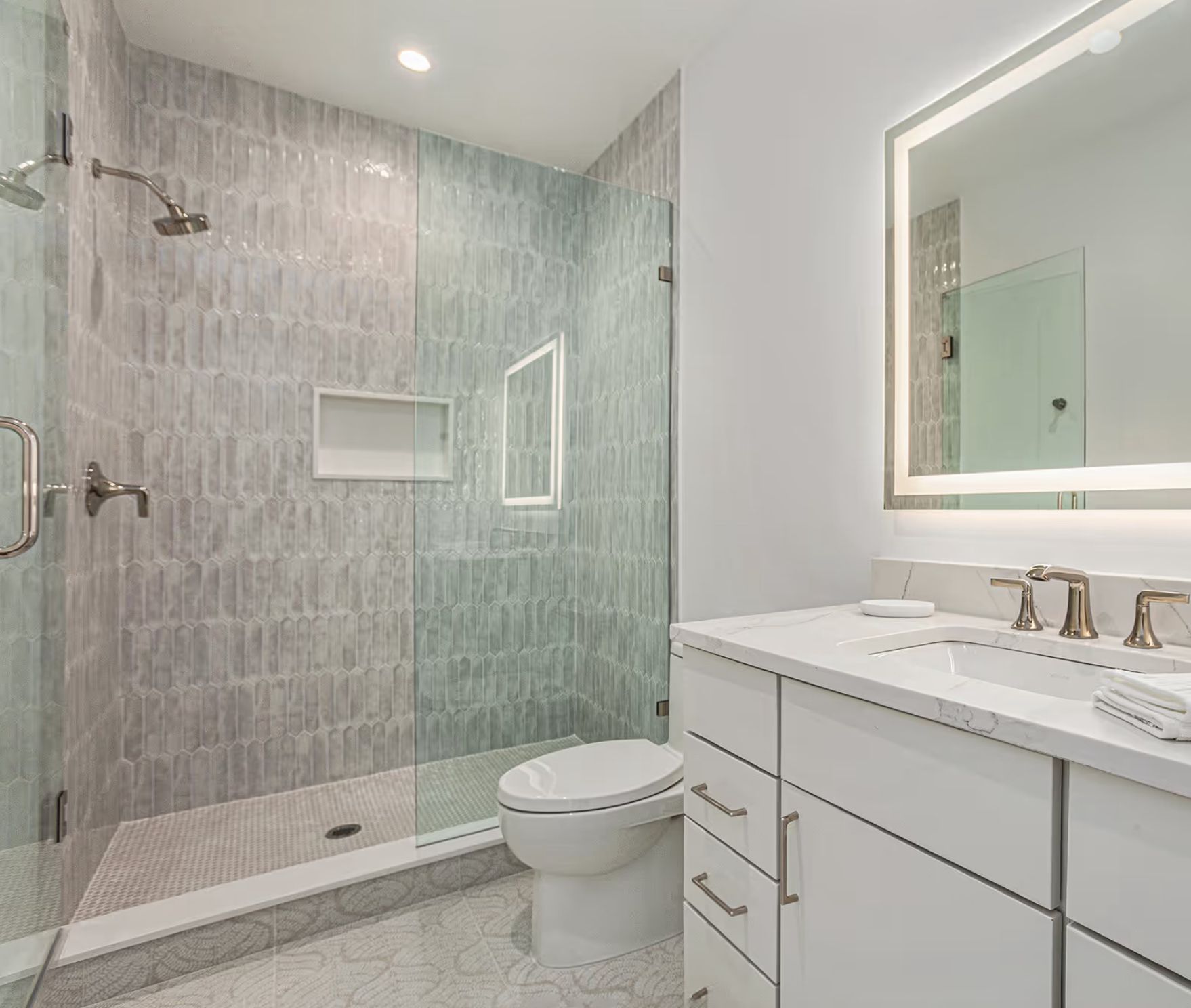 Modern bathroom with gray tiled shower, white vanity, and bright, backlit mirror