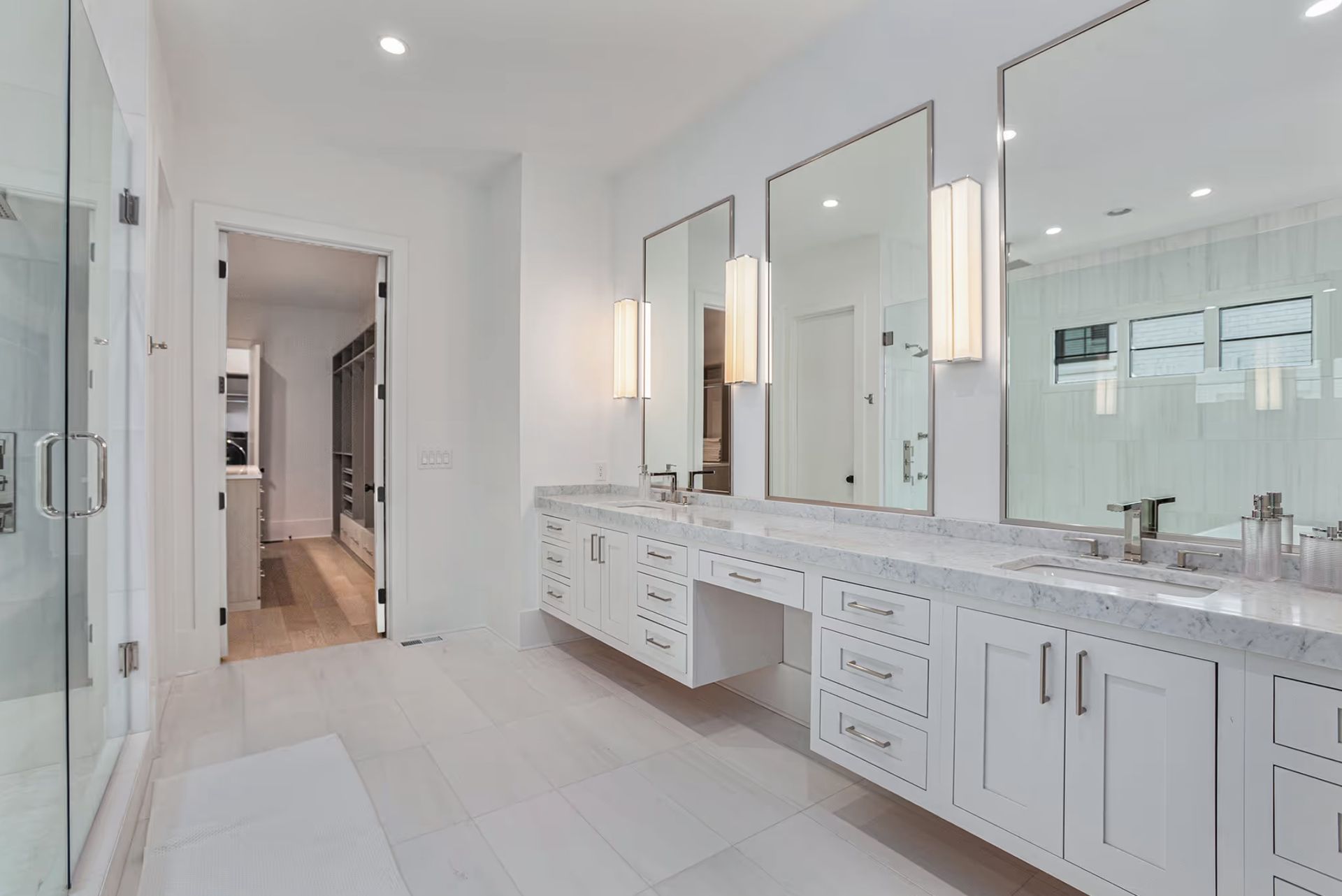 White bathroom with triple vanity, large mirrors, glass shower, and open doorway