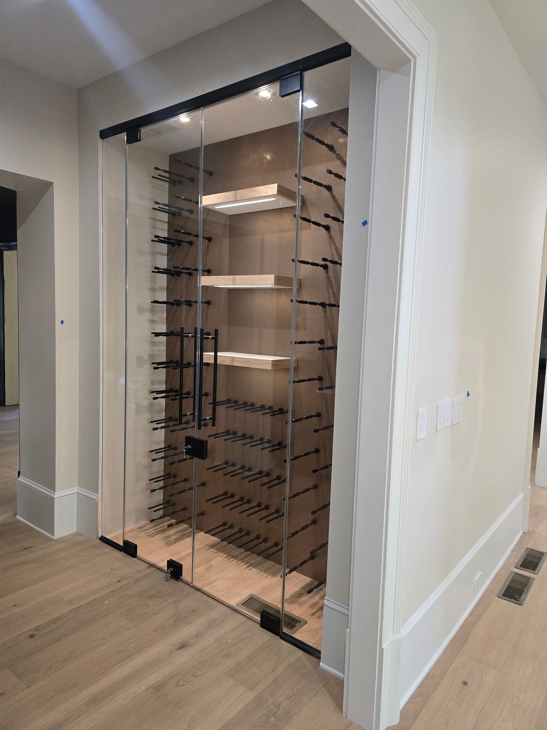 Glass-enclosed wine cellar with multiple shelves and racks for bottles, in a bright hallway