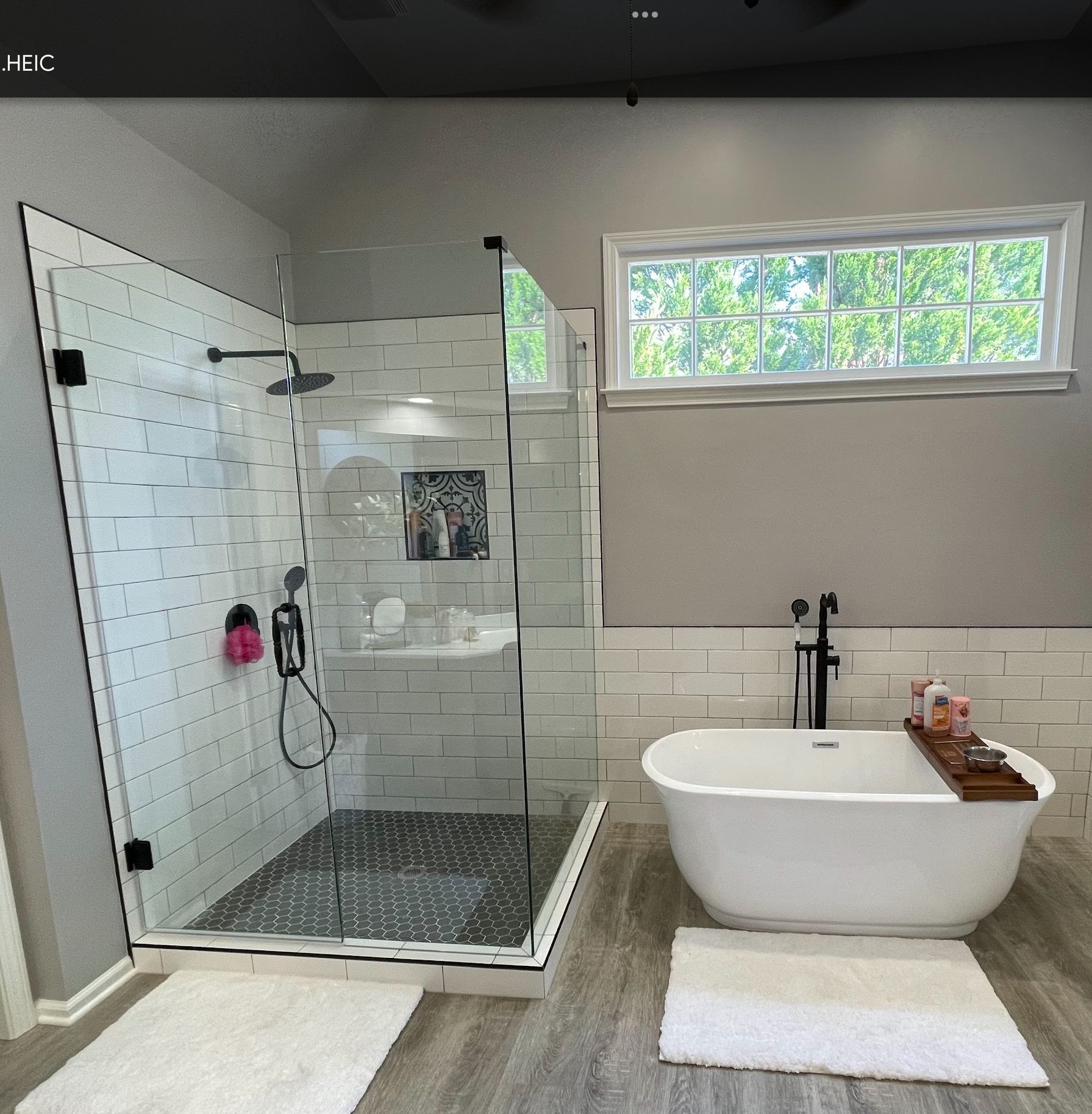 Modern bathroom: glass shower, white tub, gray walls, wooden floors, and a window.