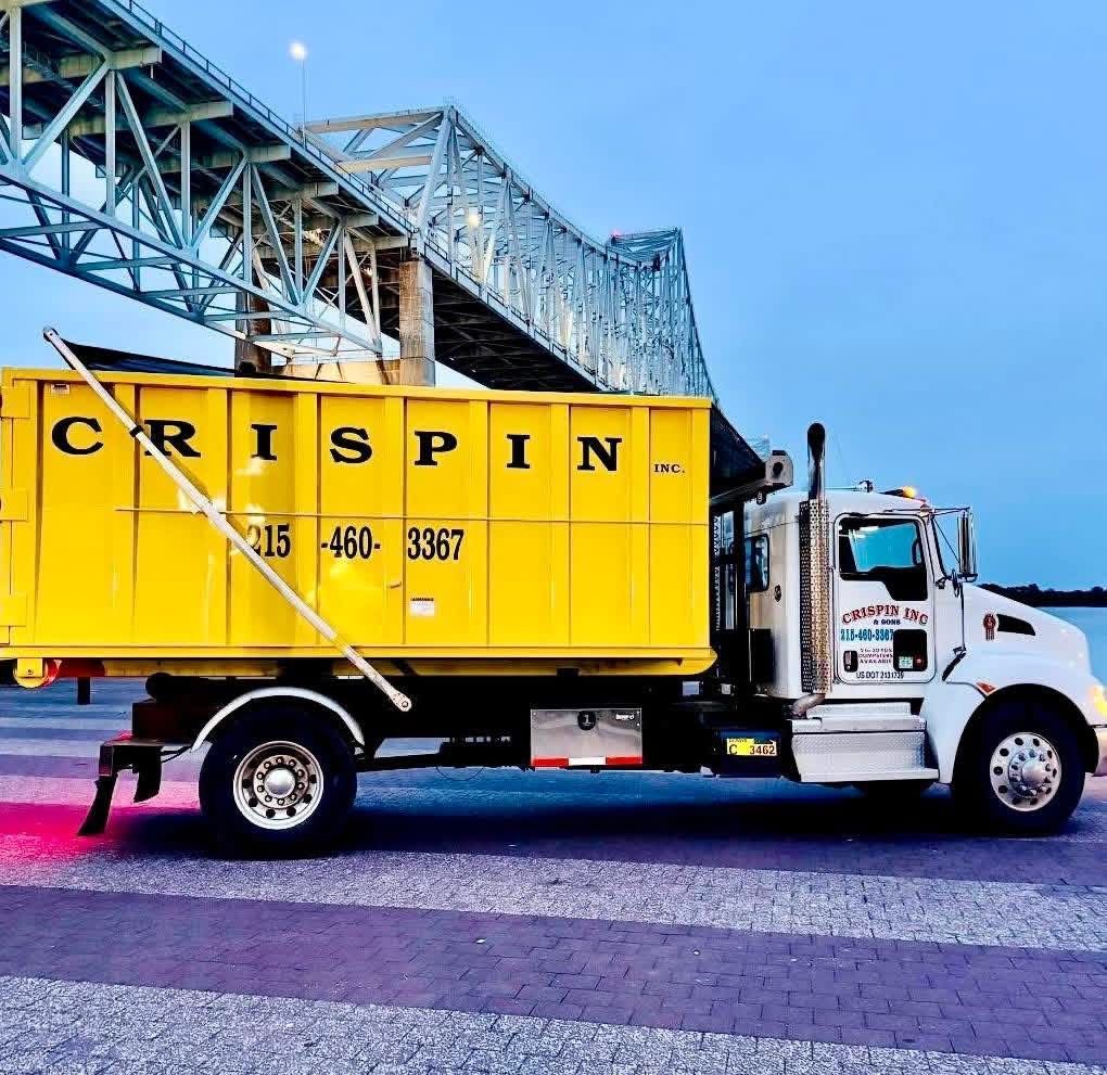 A yellow Crispin roll-off dump truck parked on a paved lot beneath a large steel bridge at dusk.