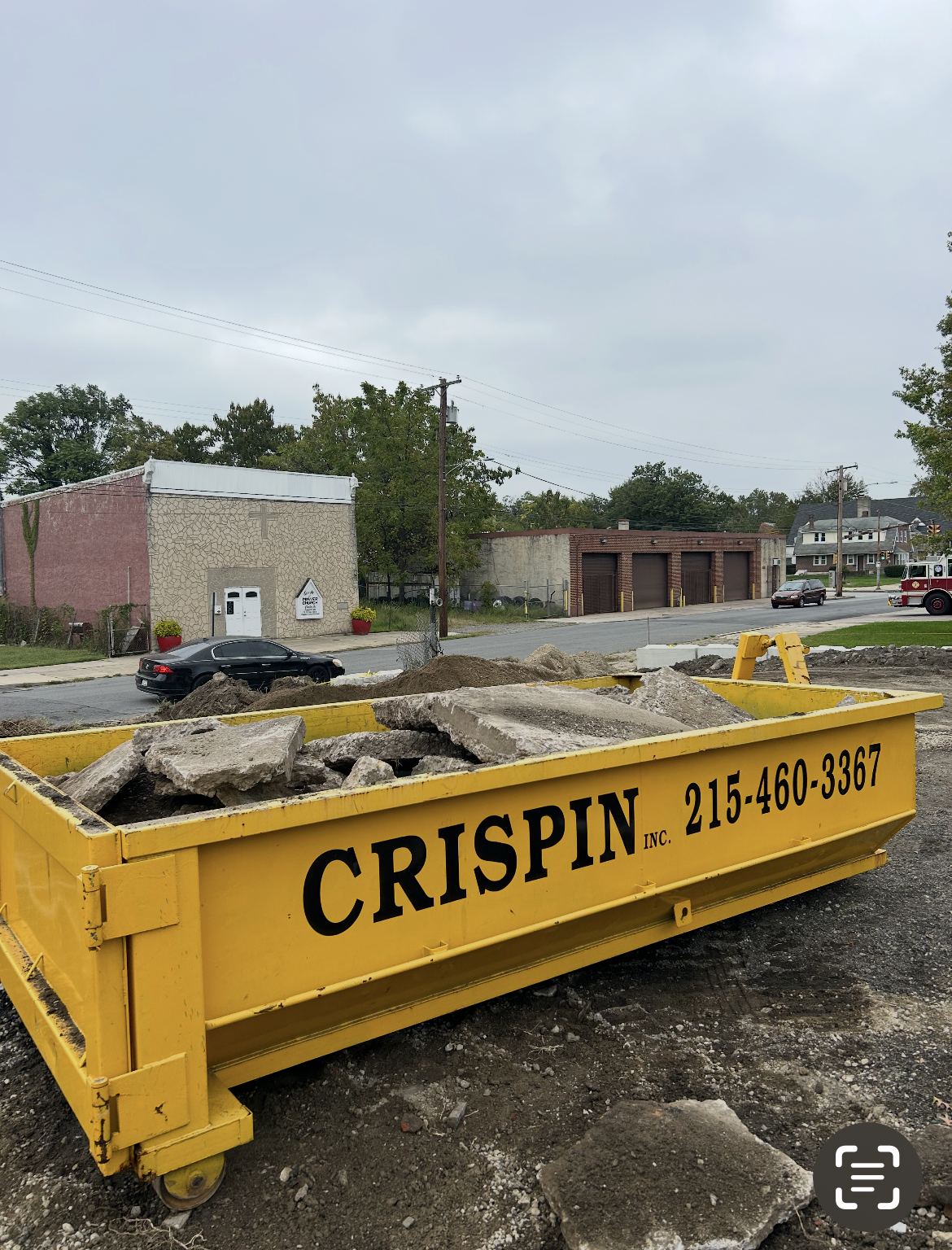 A yellow, industrial roll-off dumpster filled with concrete rubble stands on an unpaved, gravel lot near a street.