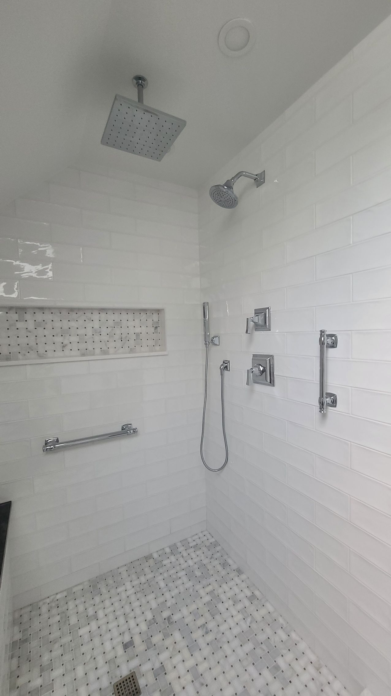 White tiled shower with rain showerhead, handheld shower, and grab bar.