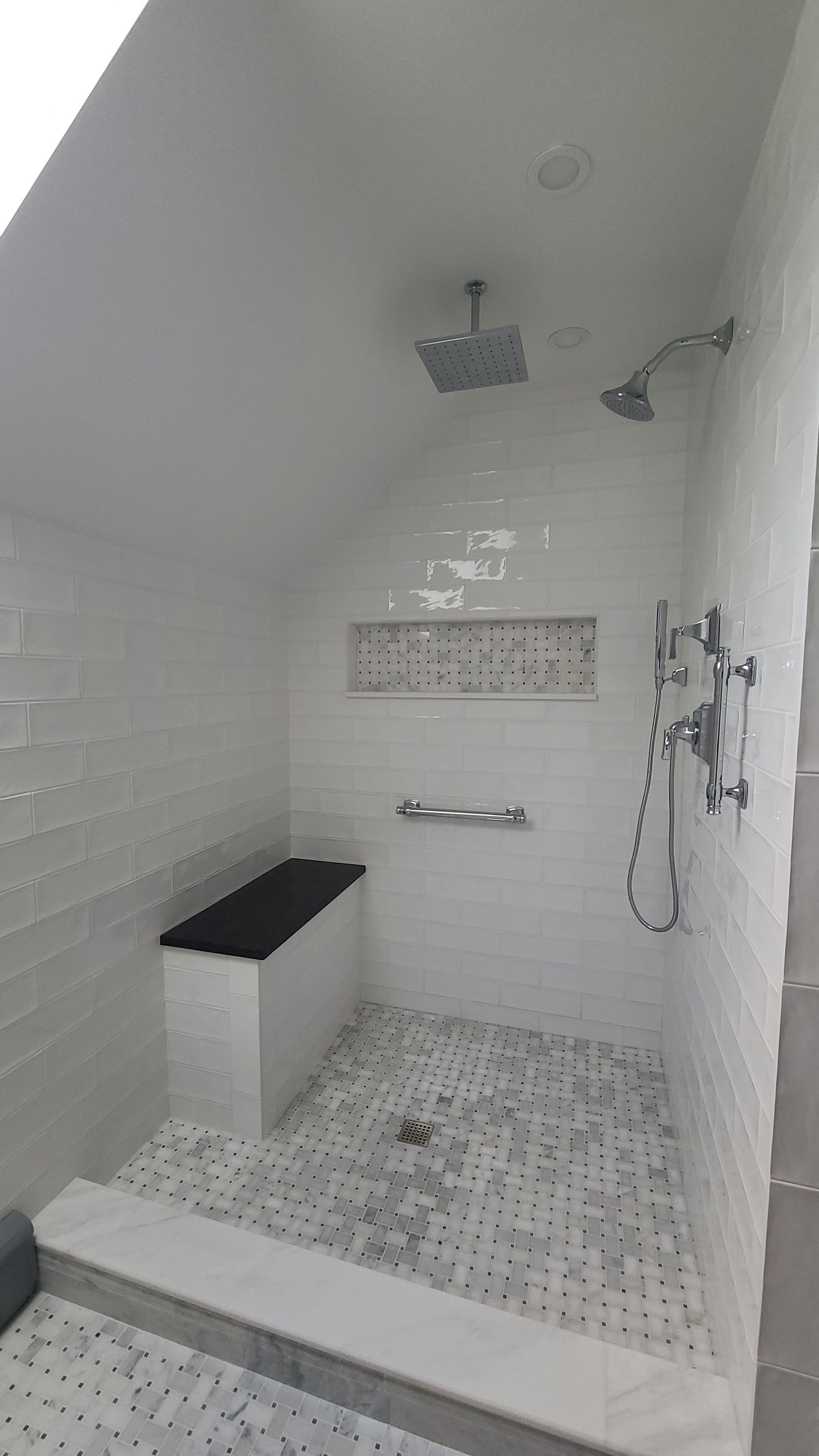 White tiled shower with built-in bench, two shower heads, grab bars, and mosaic floor tile.