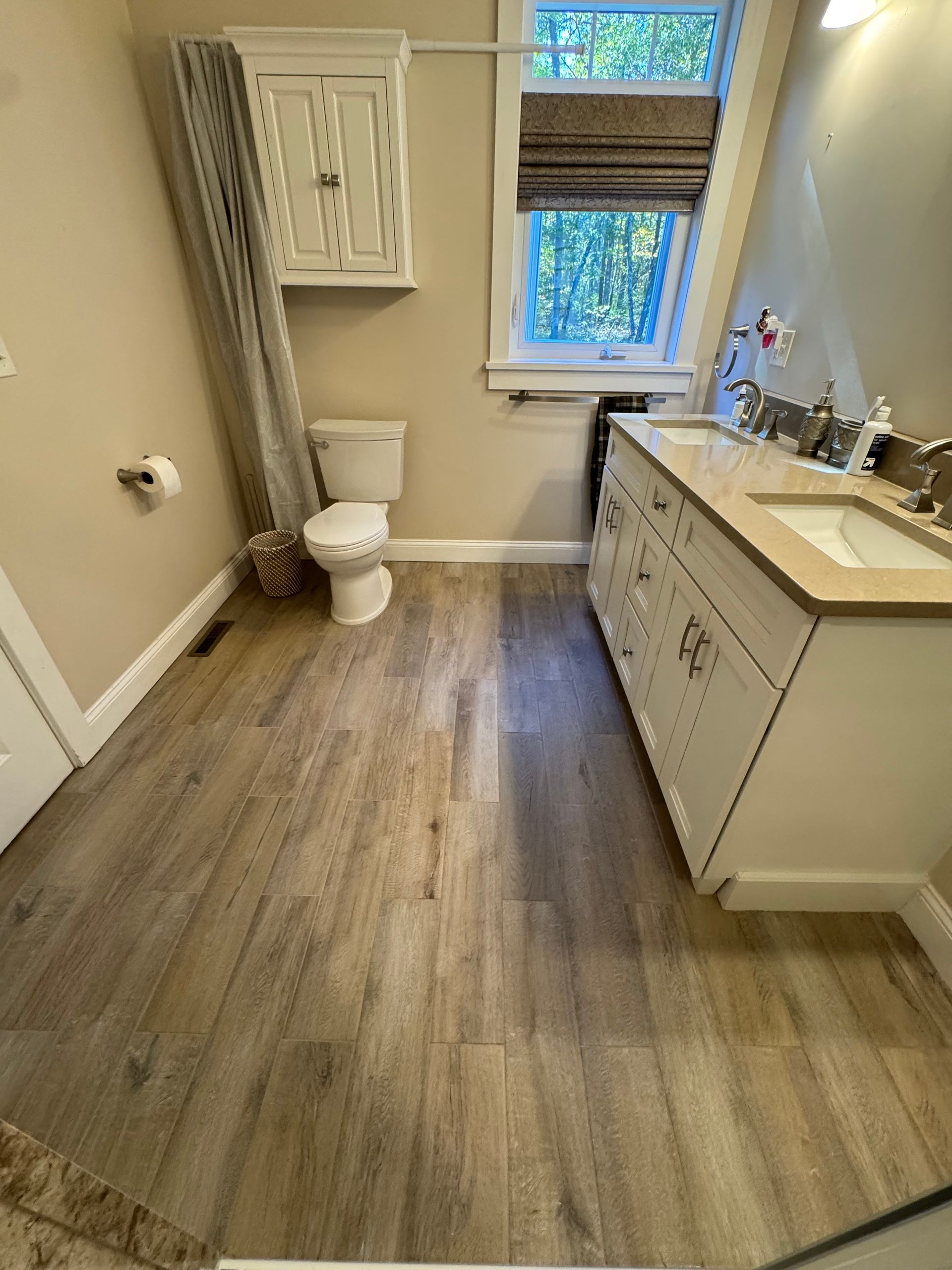 Bathroom with wood-look flooring, white vanity with double sinks, toilet, and window with shade. Light beige walls.