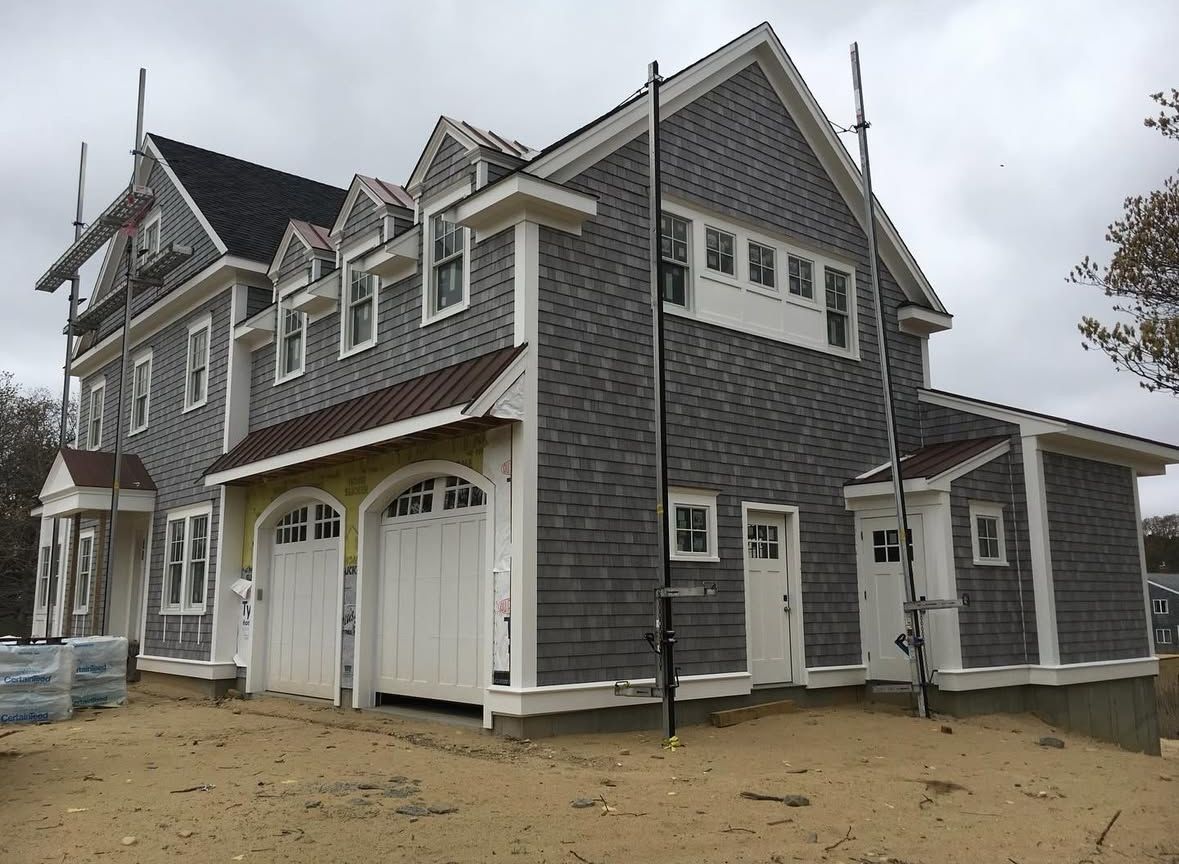 Gray shingled house with white trim, three garage doors, and multiple windows under a cloudy sky.