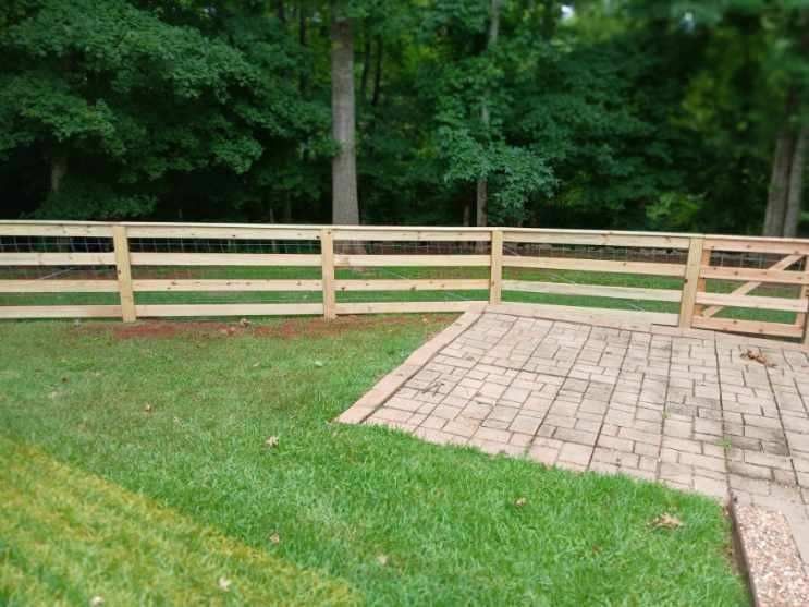 A wooden three-rail fence borders a backyard lawn next to a paver patio, with a wooded area in the background.