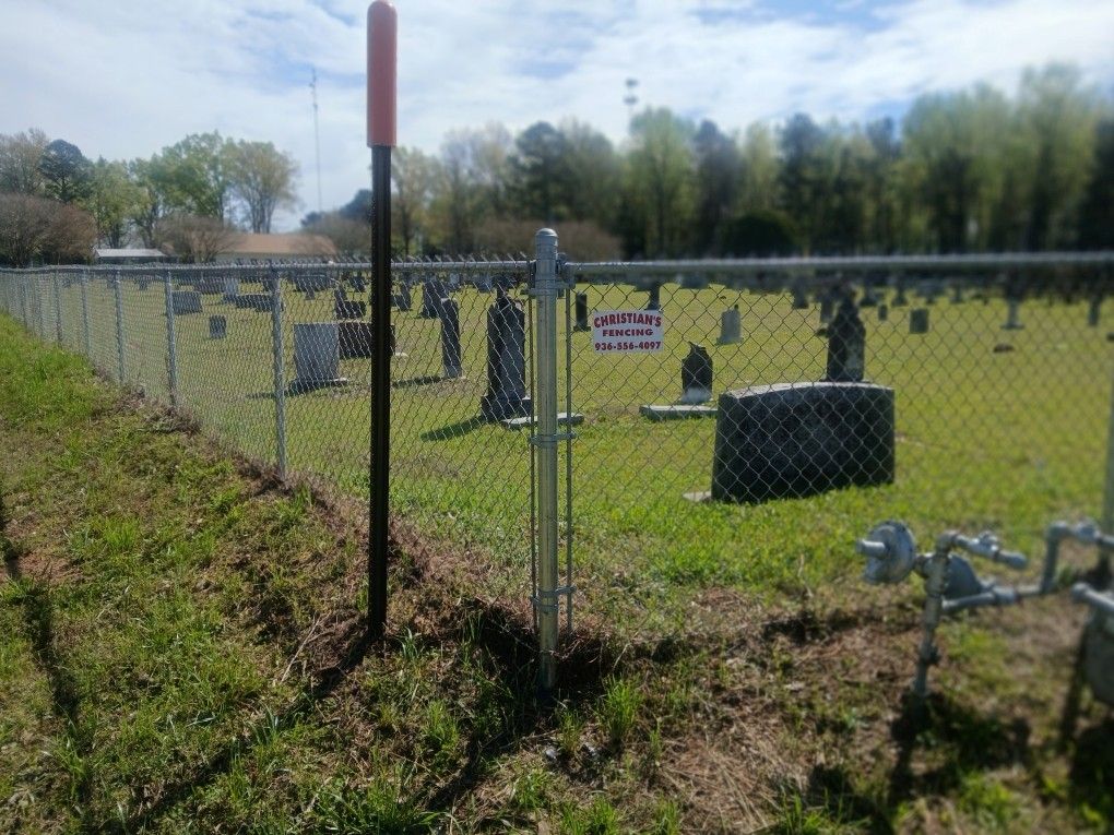 A fenced-in cemetery with rows of gravestones and an orange-topped utility marker on a sunny day.